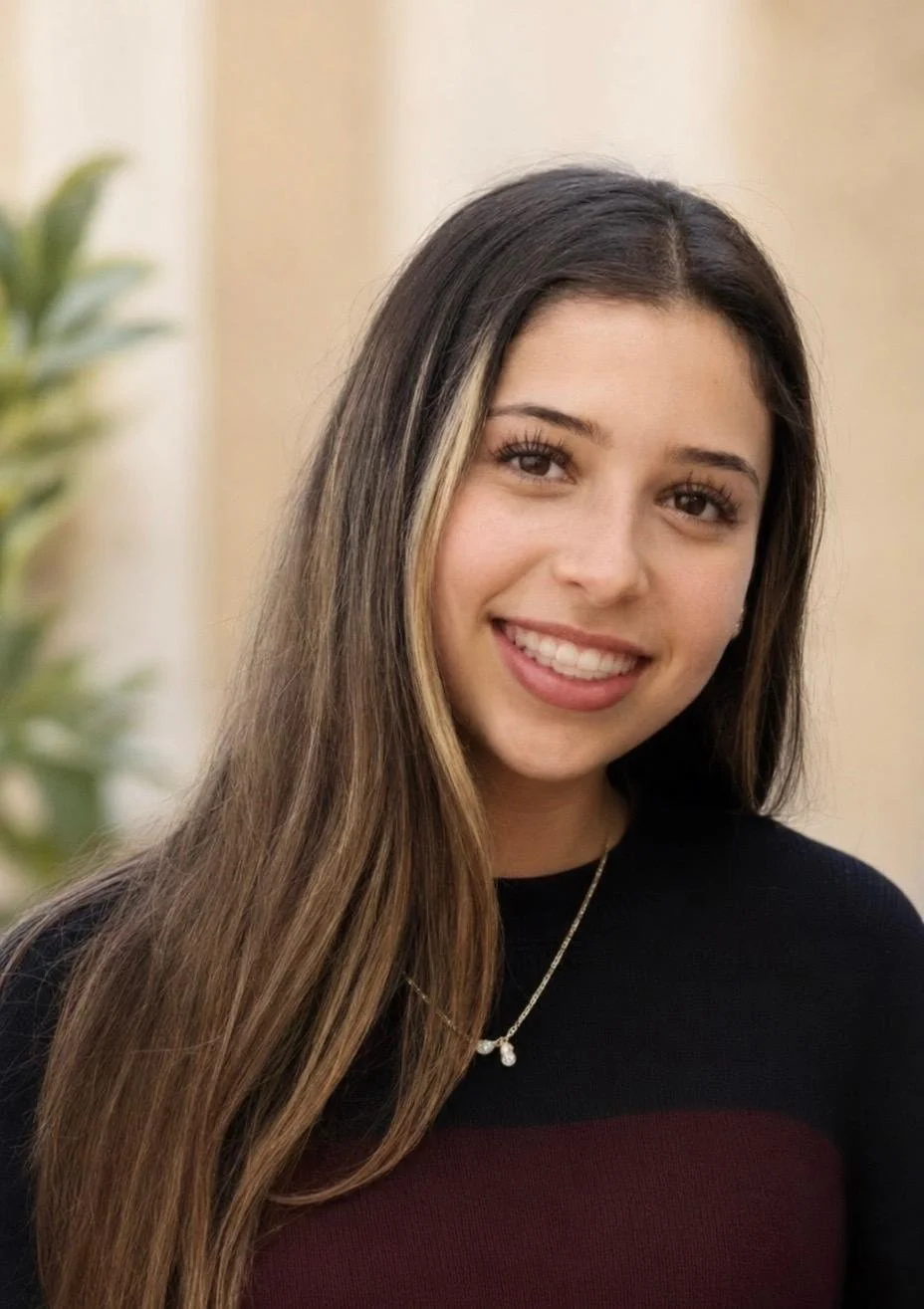 Young woman with long brown hair, smiling, wearing a black and maroon top and a pearl necklace, standing indoors with a blurred background.
