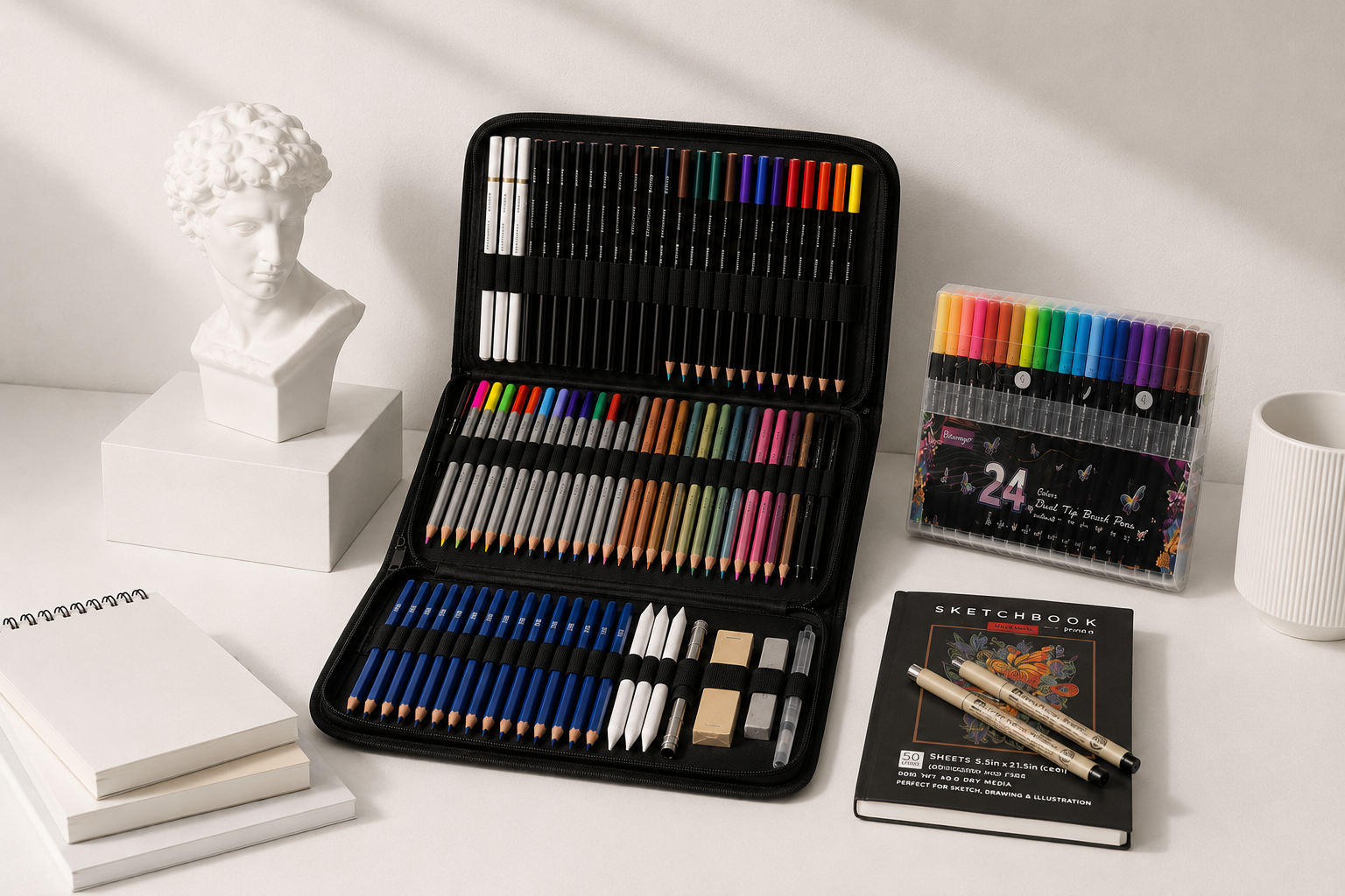 Art supplies organized on a white desk, including a closed black pencil case with colored markers, colored pencils, and drawing tools, a set of pastel brush pens, a sketchbook, two pens, a small notebook, a white bust sculpture, a stack of books or notebooks, and a white textured cup.