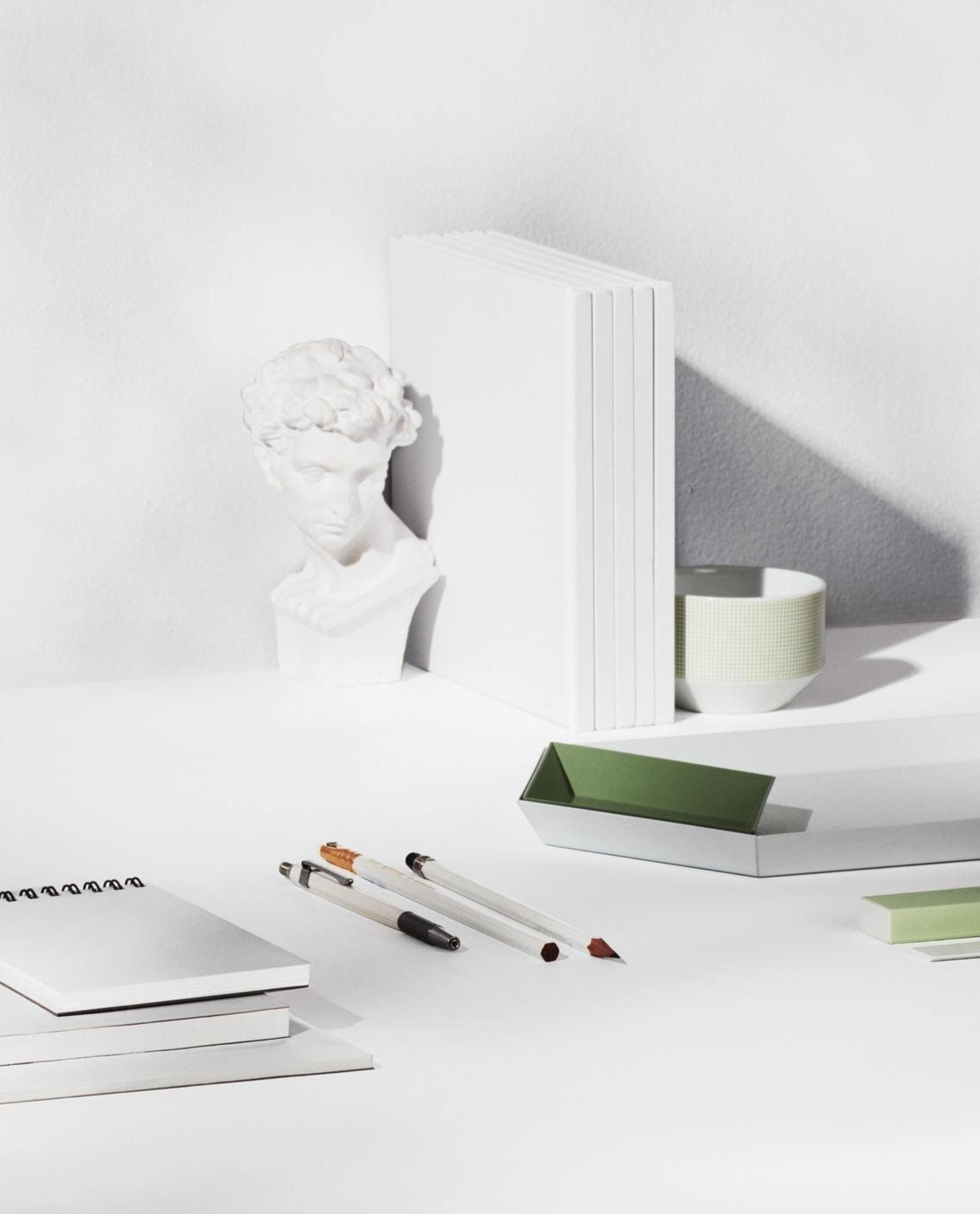 Minimalist workspace with a white bust sculpture, white books, a green and white bowl, a green tray, notebooks, pens, and pencils on a white desk.