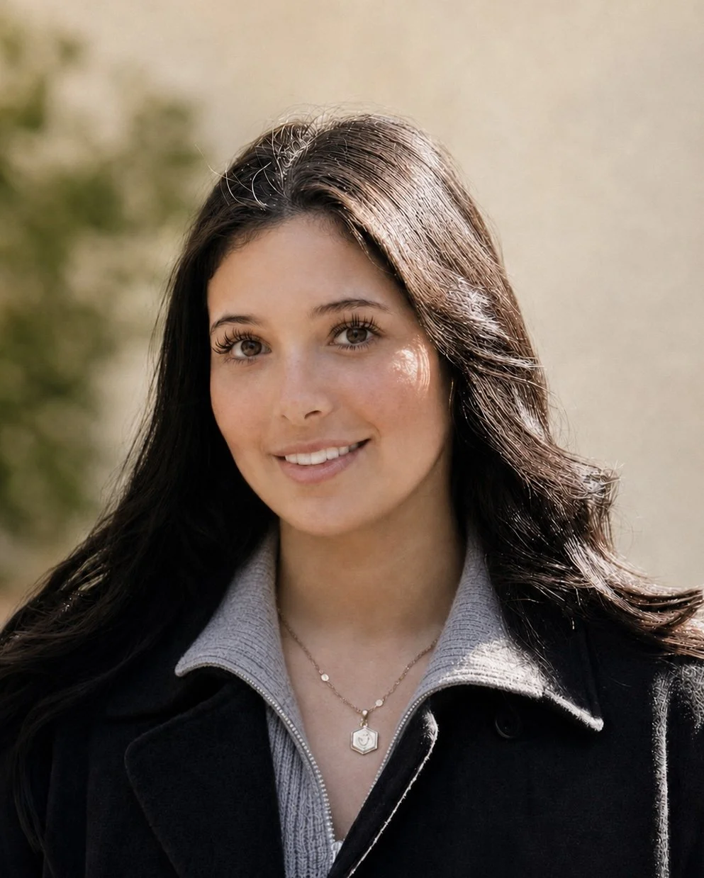 A young woman with long dark hair and brown eyes smiling outdoors, wearing a gray zip-up jacket with a collar and a gold necklace with a pendant, with blurred greenery in the background.