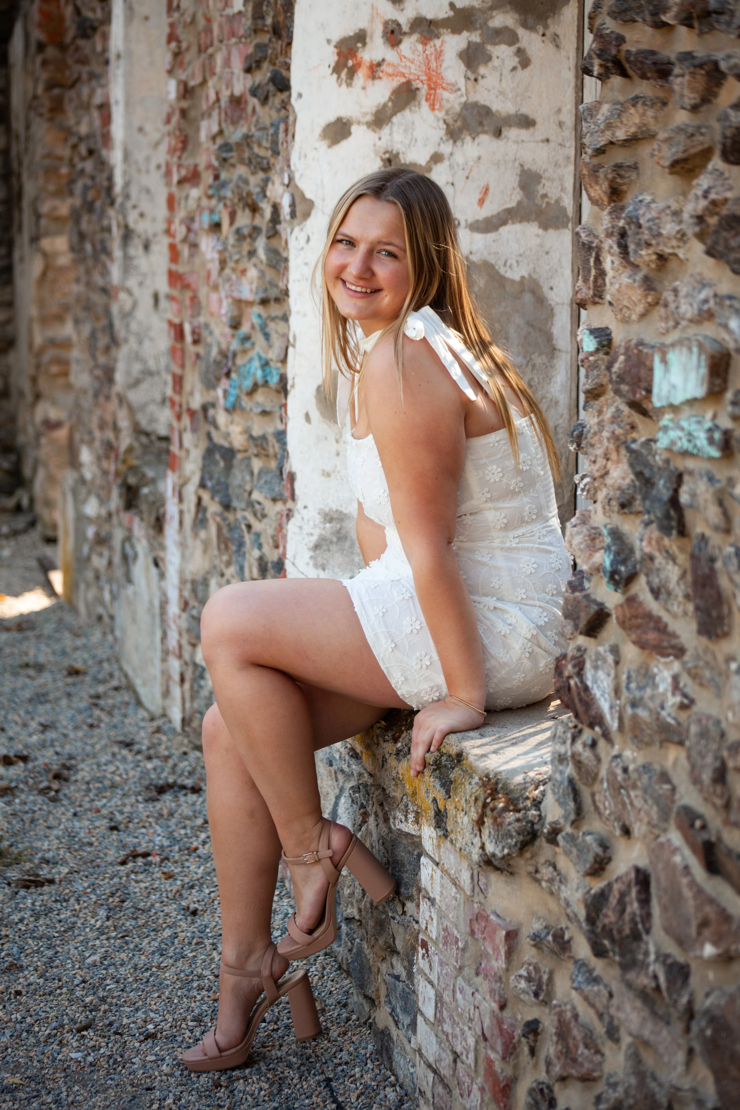 Senior portrait of a young woman in a white lace dress sitting on a stone ledge at a rustic ruins location, smiling over her shoulder at the camera.