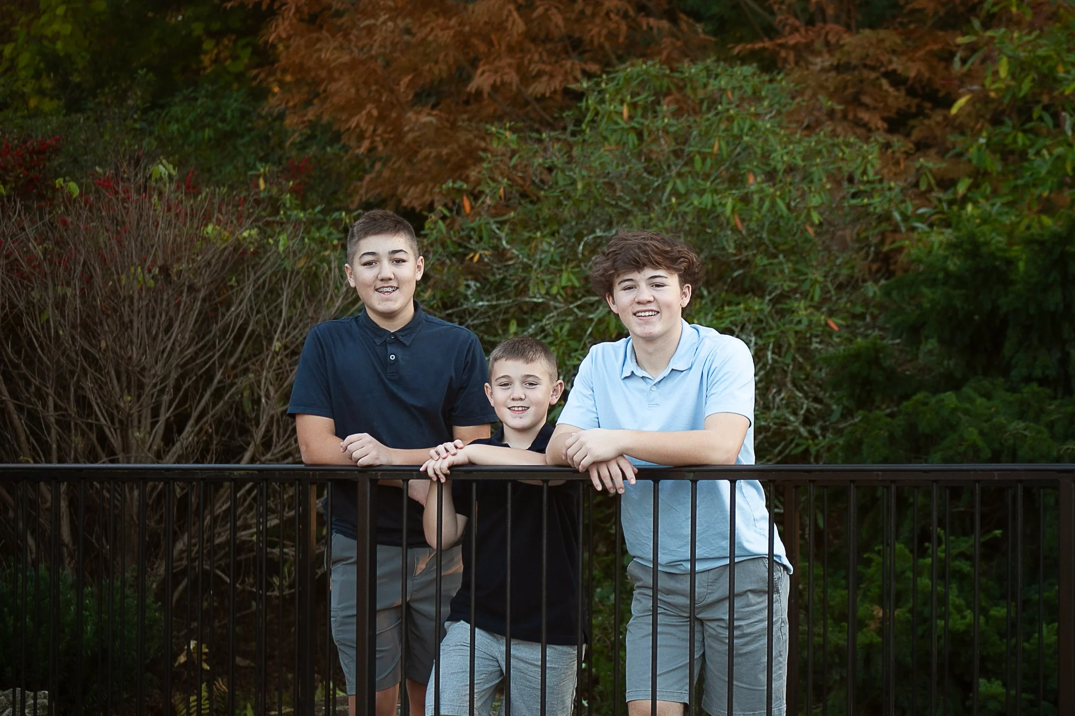 Three brothers leaning on a railing surrounded by fall foliage, candid sibling portrait Chester County PA