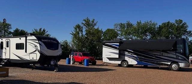 A recreational vehicle (RV) and a red pickup truck parked on a gravel lot with trees in the background.