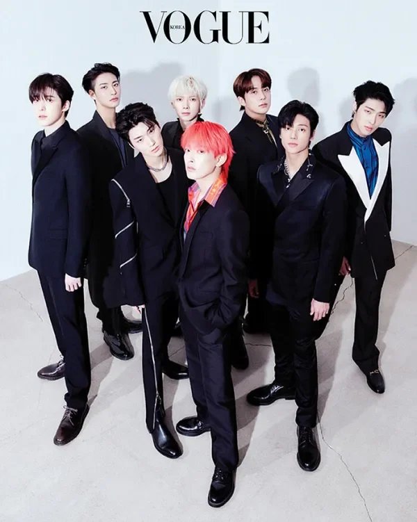 A seven-member K-pop boy band posing for a Vogue photo shoot, dressed in stylish black suits, standing in front of a white wall with the Vogue logo at the top.