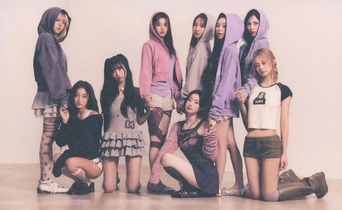 A group of nine young women posing together against a plain wall. They are dressed in colorful casual and sporty outfits, some wearing hoodies and others in skirts and shorts, with a variety of hairstyles and expressions.