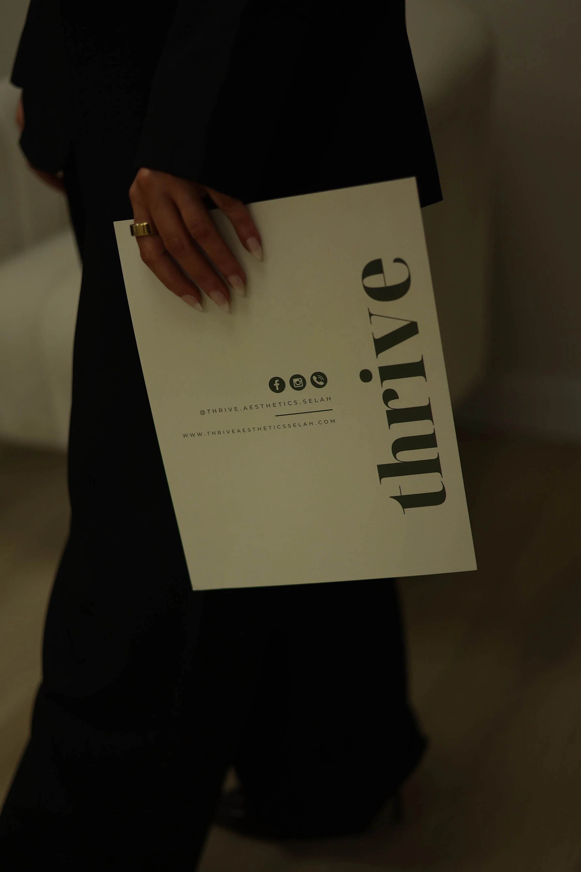 Person wearing black clothing holding a white paper with the words 'LOVE.' printed vertically, and social media icons with the handle '@THRIVE.AESTHETICS.SELAH' and website 'WWW.THRIVEAESTHETICSELAH.COM'.
