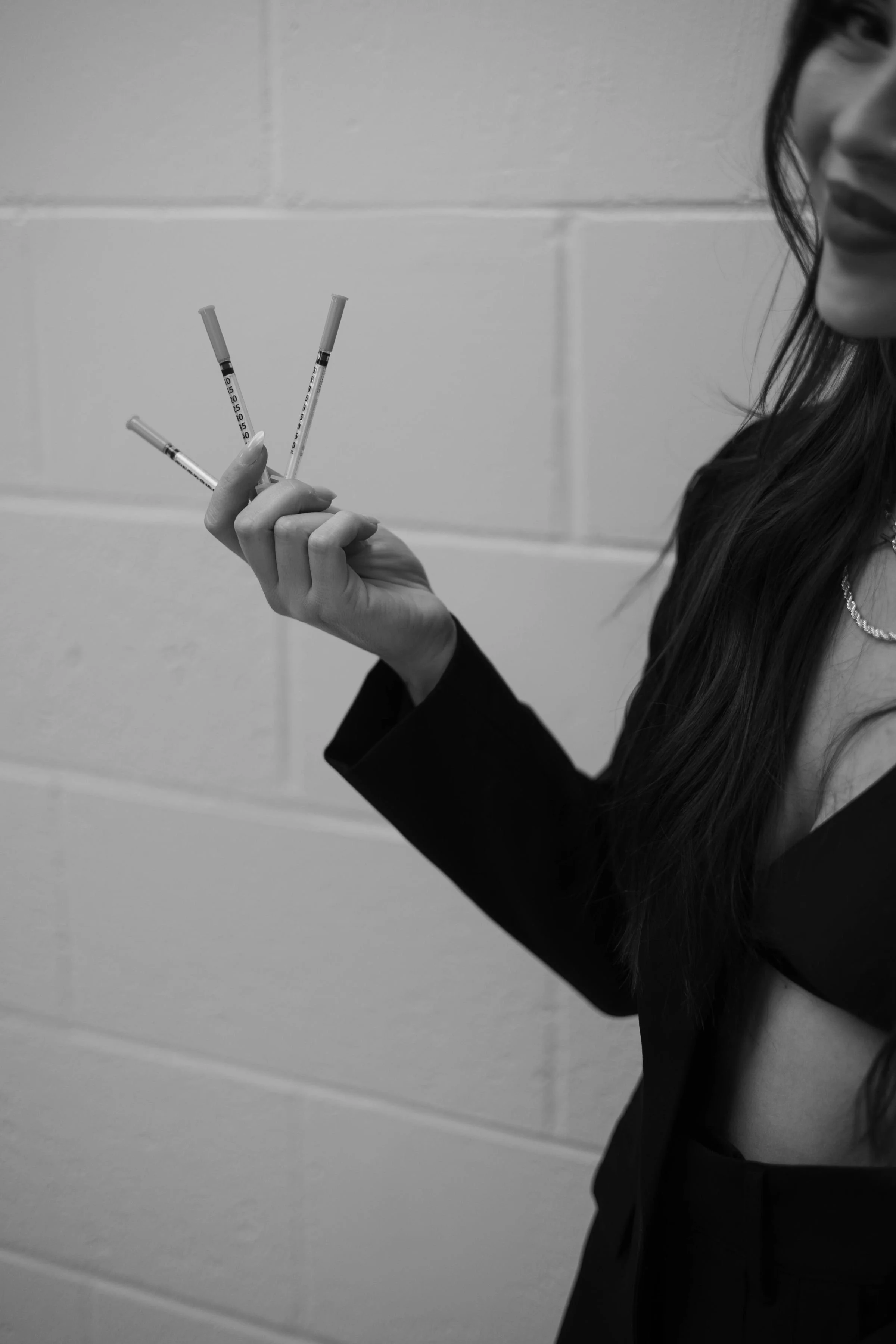 Black and white photo of a woman holding three syringes, with her face partially visible on the right side of the image, against a plain wall background.
