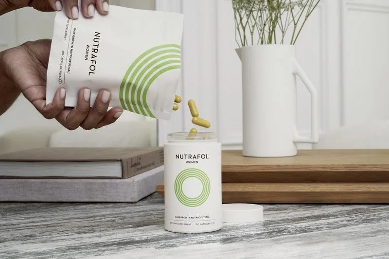 A hand pouring yellow capsules from a white packet labeled 'NUTRAFOL Women' into a white container on a wooden surface, with a white vase holding green plants in the background.