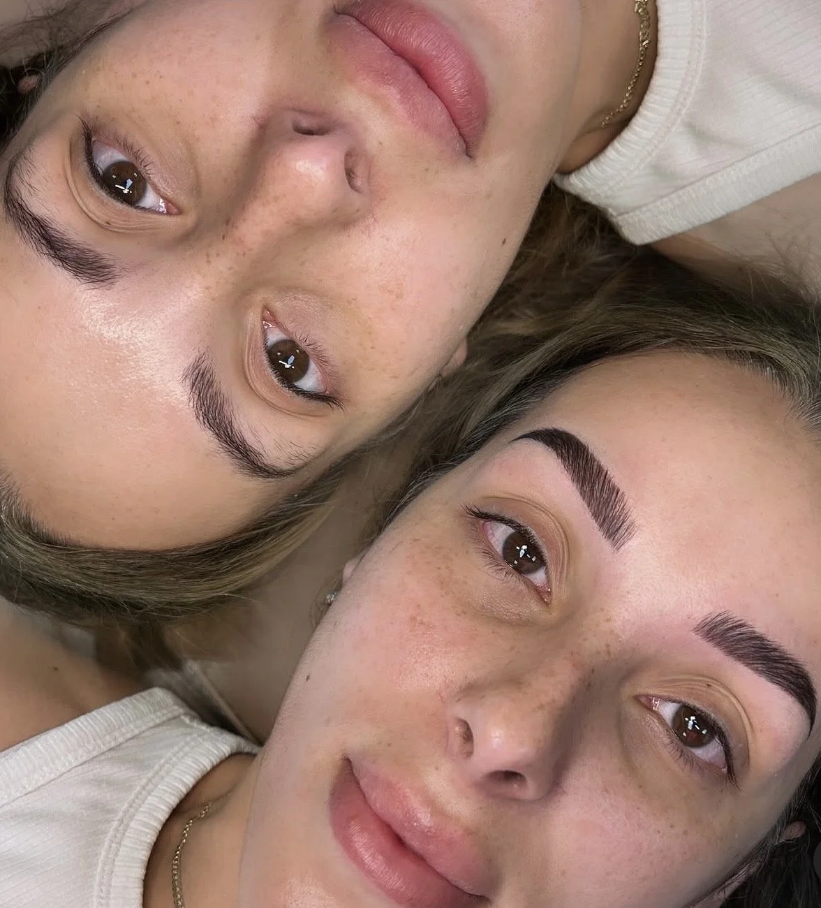 Two women lying side by side, showing their faces with bright makeup, one with a natural look and the other with bold eyebrows and subtle makeup.