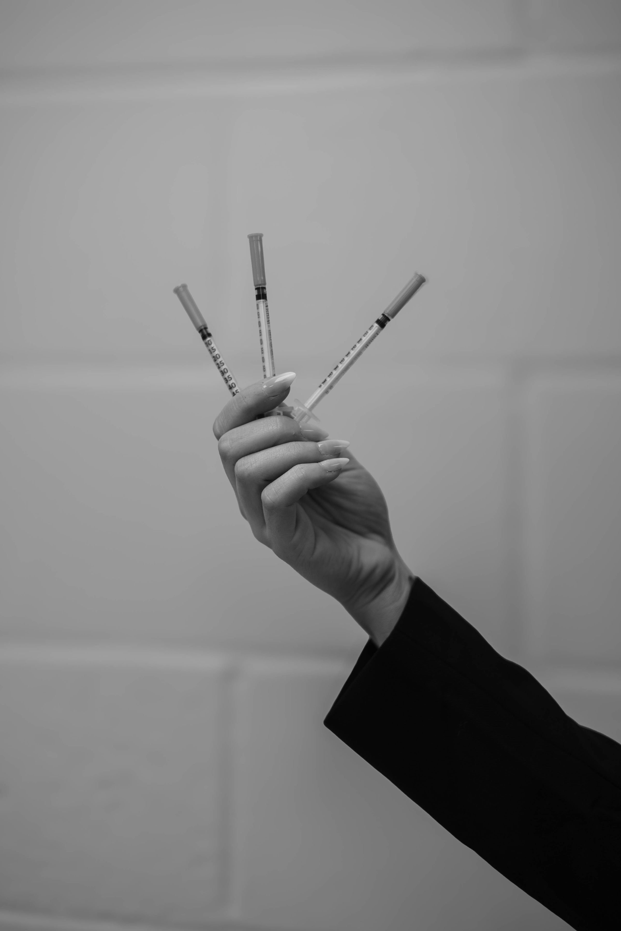 A person in a black suit holding four syringes with their hand in front of a plain background.
