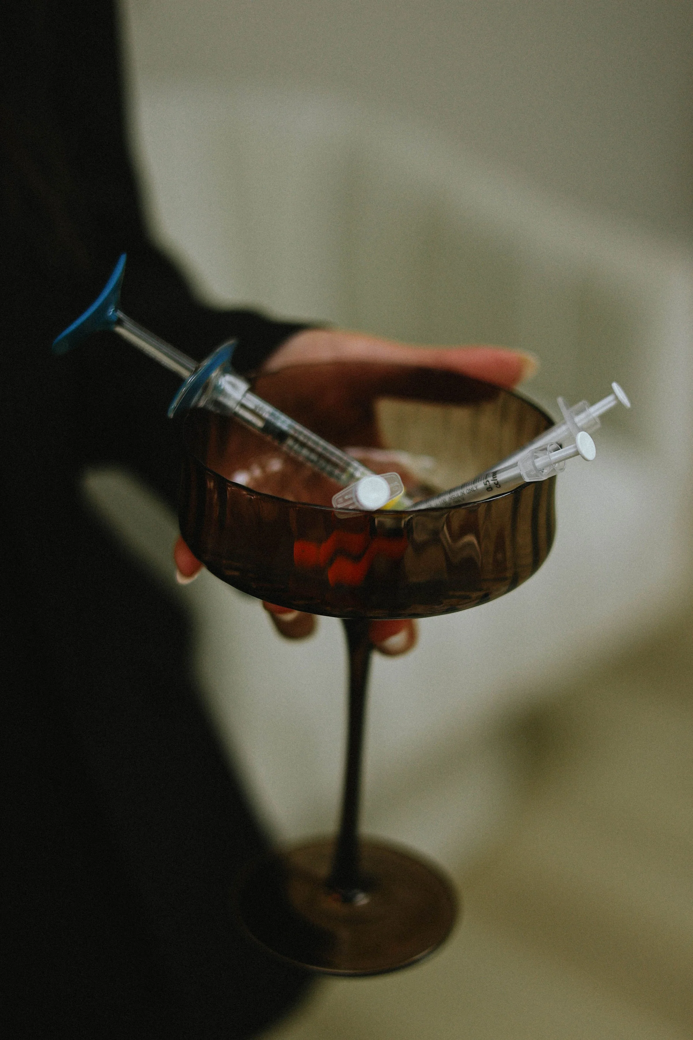 A person holding a brown glass with blood dripping from the rim, a syringe filled with liquid is in the person's hand.