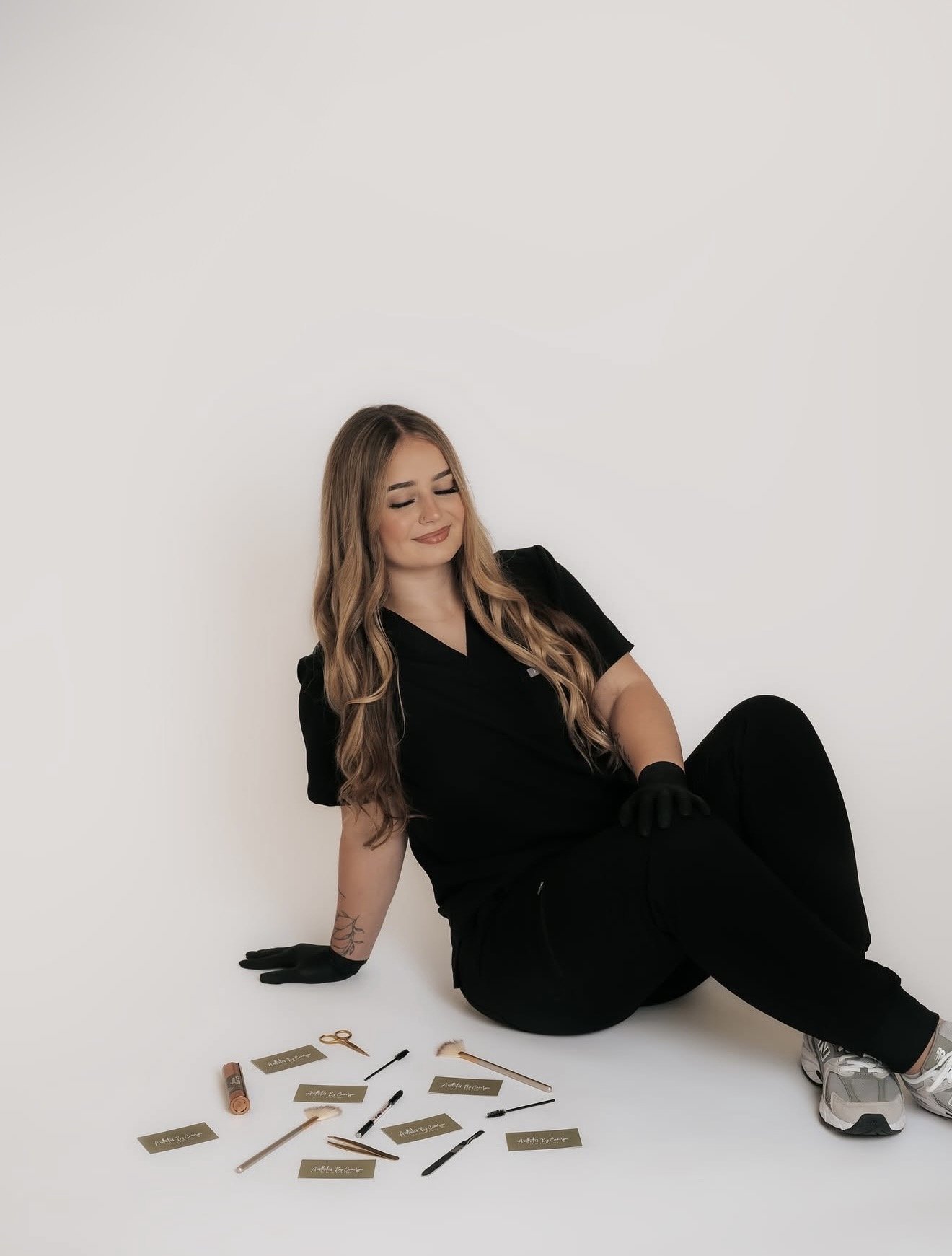 A woman dressed in black, sitting on the floor with her eyes closed and smiling, surrounded by makeup tools and name tags, posing against a plain white background.