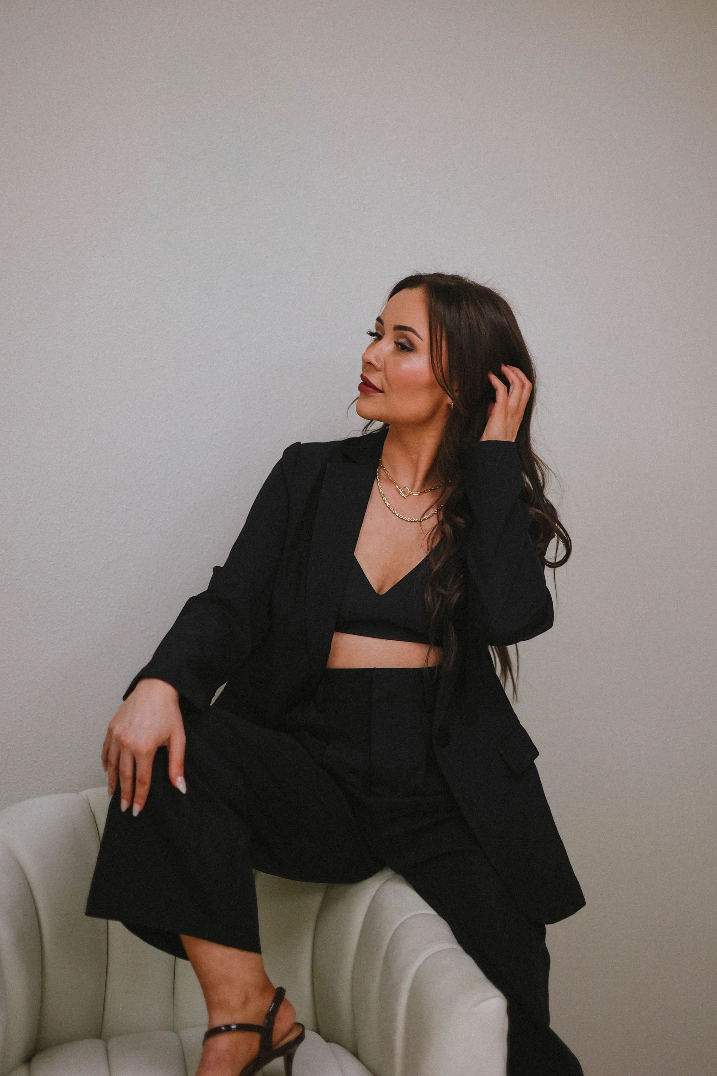 A woman with long dark hair, wearing a black blazer and matching pants, sitting on a white tufted armchair against a plain wall, looking to her left, with one hand touching her hair and the other resting on her knee.