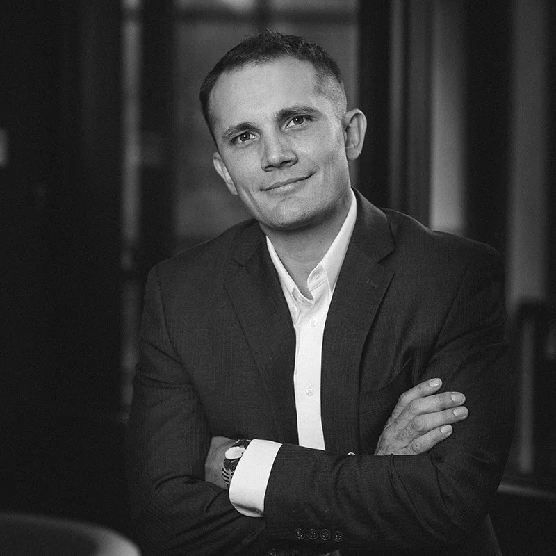 Black and white photo of a man in a suit with arms crossed, smiling, in an indoor setting.