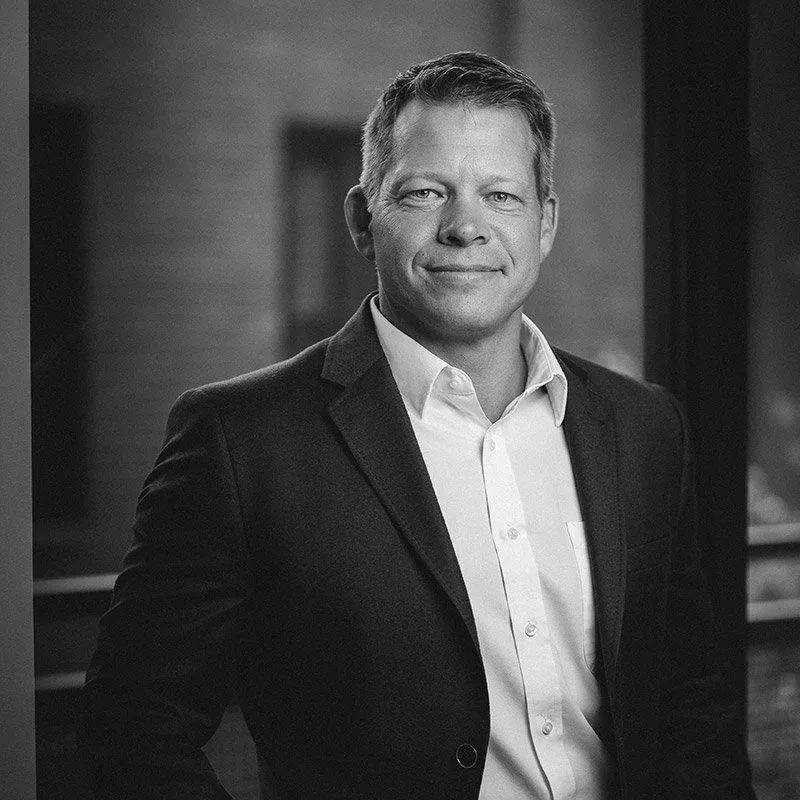 Black-and-white portrait of a man wearing a suit and white shirt, smiling slightly, standing indoors.