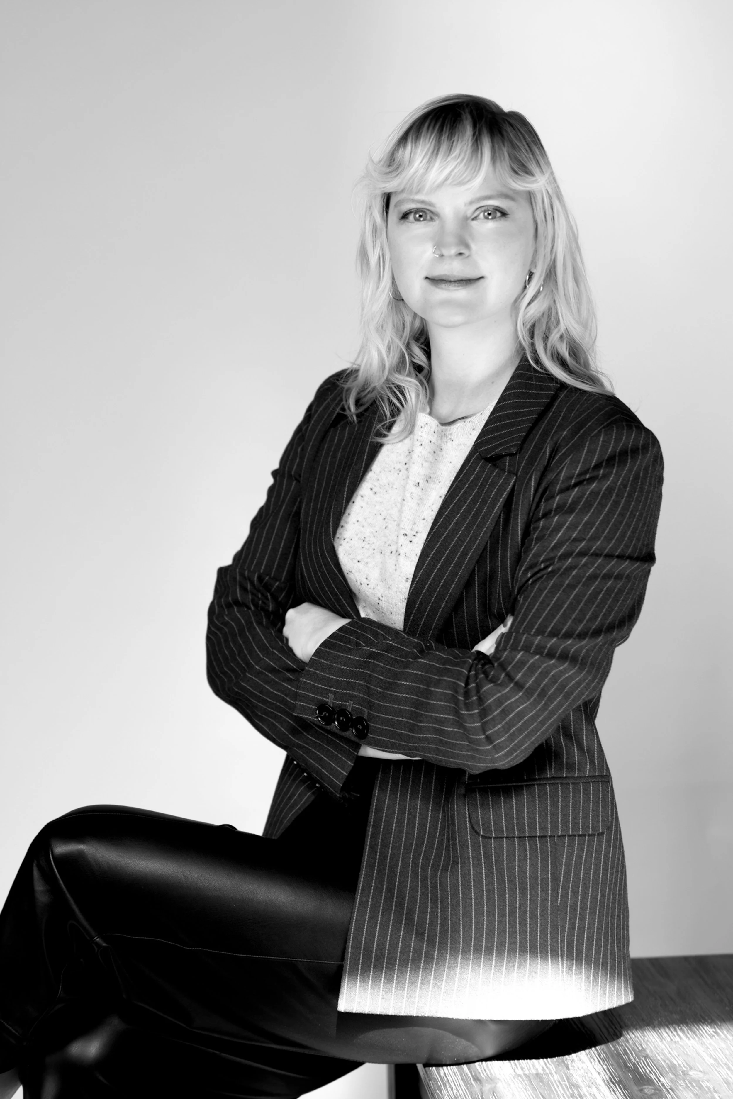 Black and white photo of a woman with wavy blonde hair, wearing a striped blazer and light-colored top, sitting with her arms crossed on a chair, against a plain background.