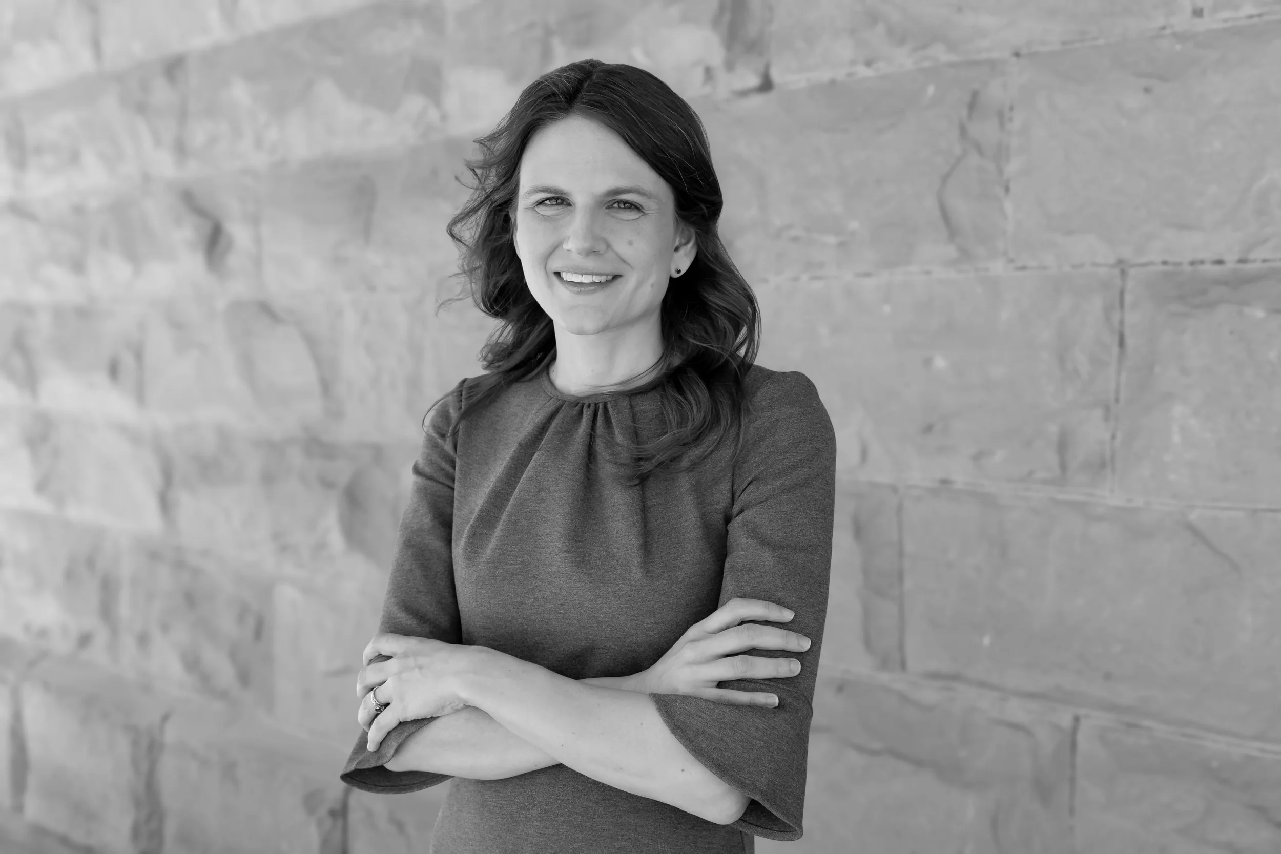 A black and white portrait of a woman with shoulder-length dark hair, standing with her arms crossed in front of a stone wall.