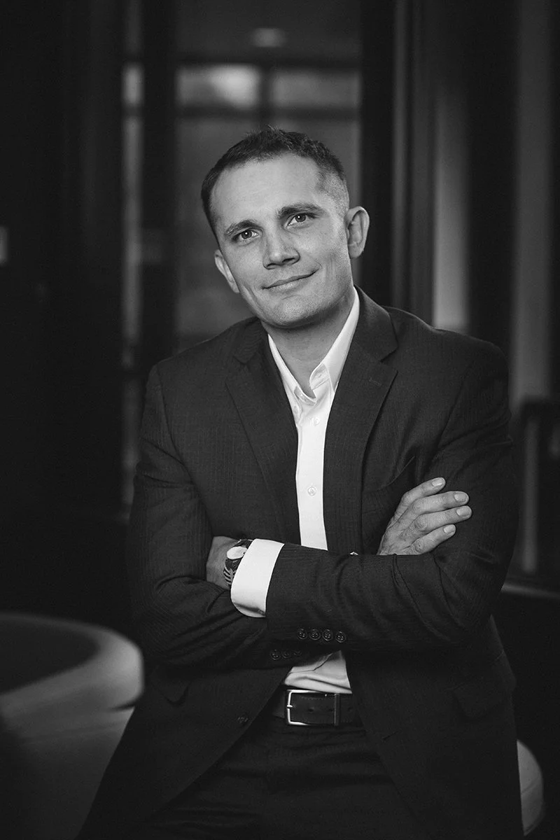 Black and white portrait of a man wearing a suit with arms crossed, standing in front of a window.