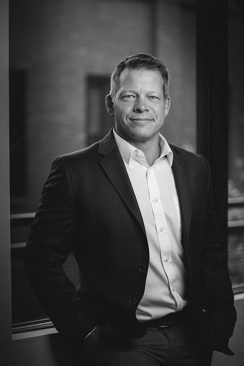 Black and white portrait of a man in a suit smiling, standing indoors near a window.