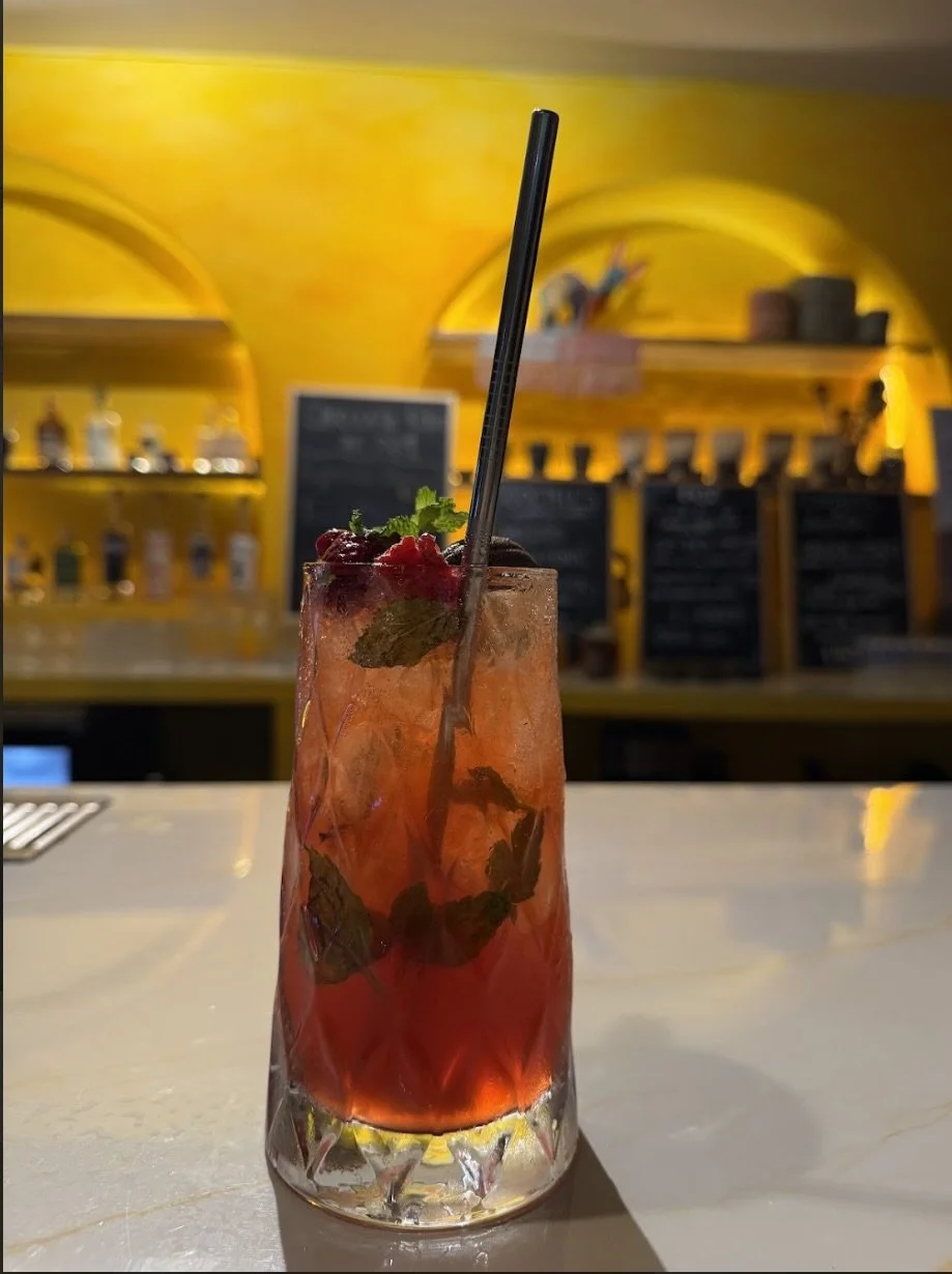A tall glass of a raspberry mojito cocktail with mint leaves, berries, a black straw, and ice cubes on a bar counter, with a yellow arched wall and shelves in the background.
