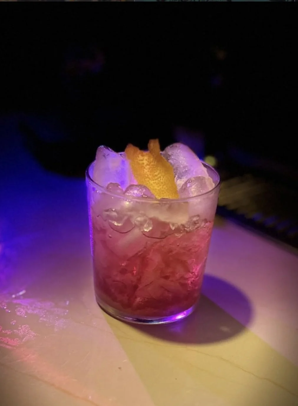 An old fashioned cocktail garnished with a lemon peel, served on a table with purple and yellow lighting.