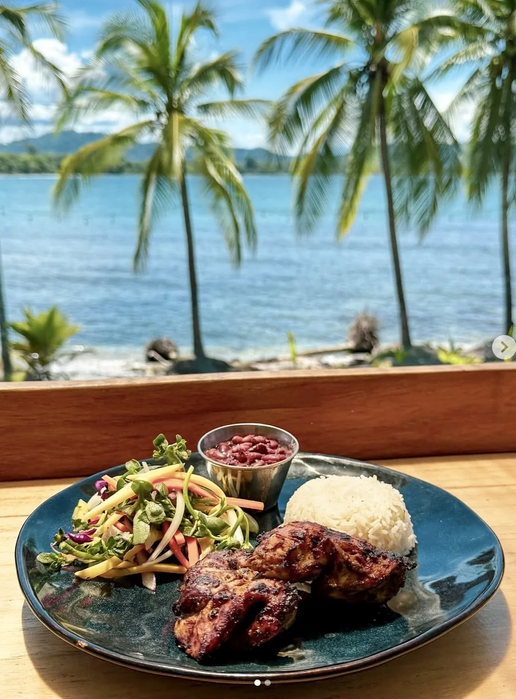 A plate of grilled chicken, rice, a colorful salad, and a small cup of beans served on a wooden table with a beach view of palm trees and the ocean in the background.
