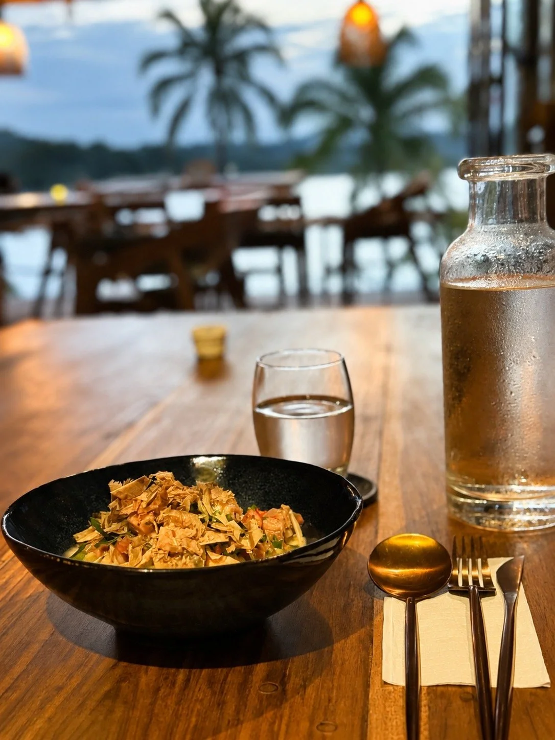 A dining table set with a bowl of Peruvian Ceviche, a glass of water, a water pitcher, and cutlery in a restaurant with a view of trees and water outside.
