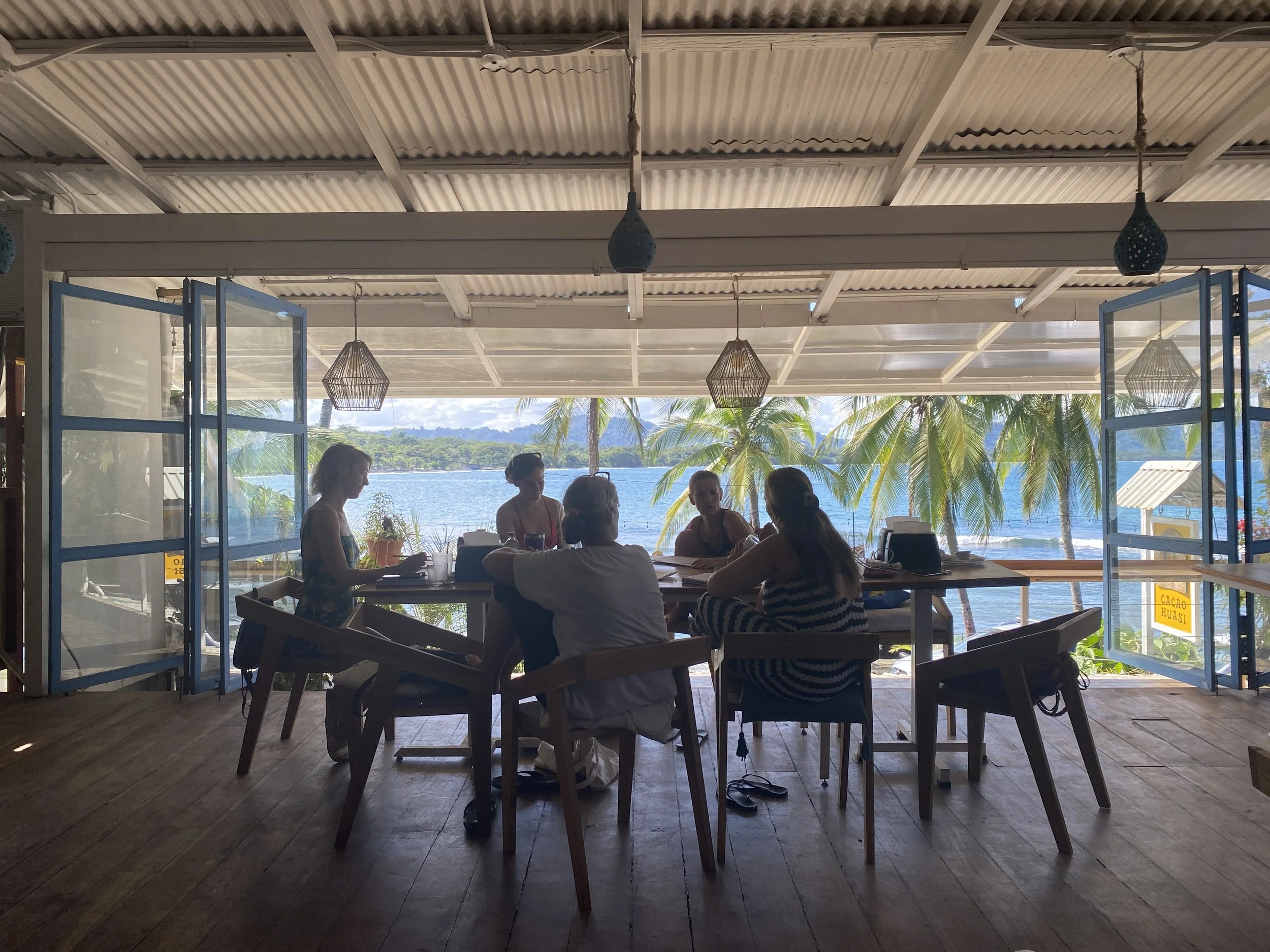 People sitting at a wooden table inside a beachside restaurant with open blue-framed glass doors, overlooking palm trees, water, and a cloudy sky.
