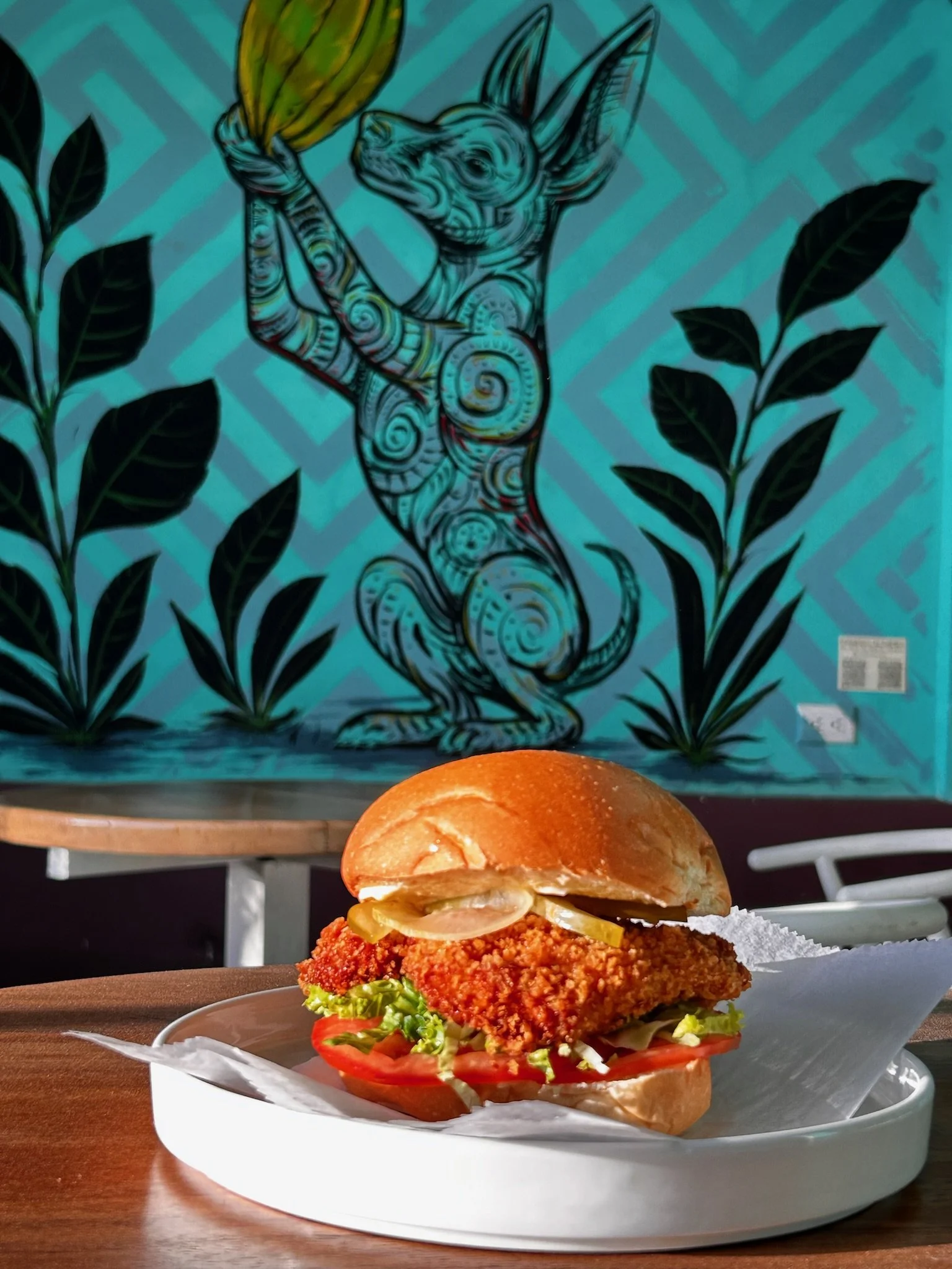 A crispy chicken sandwich with lettuce, tomato, pickles, and sauce in a bun, served on a white plate on a wooden table.