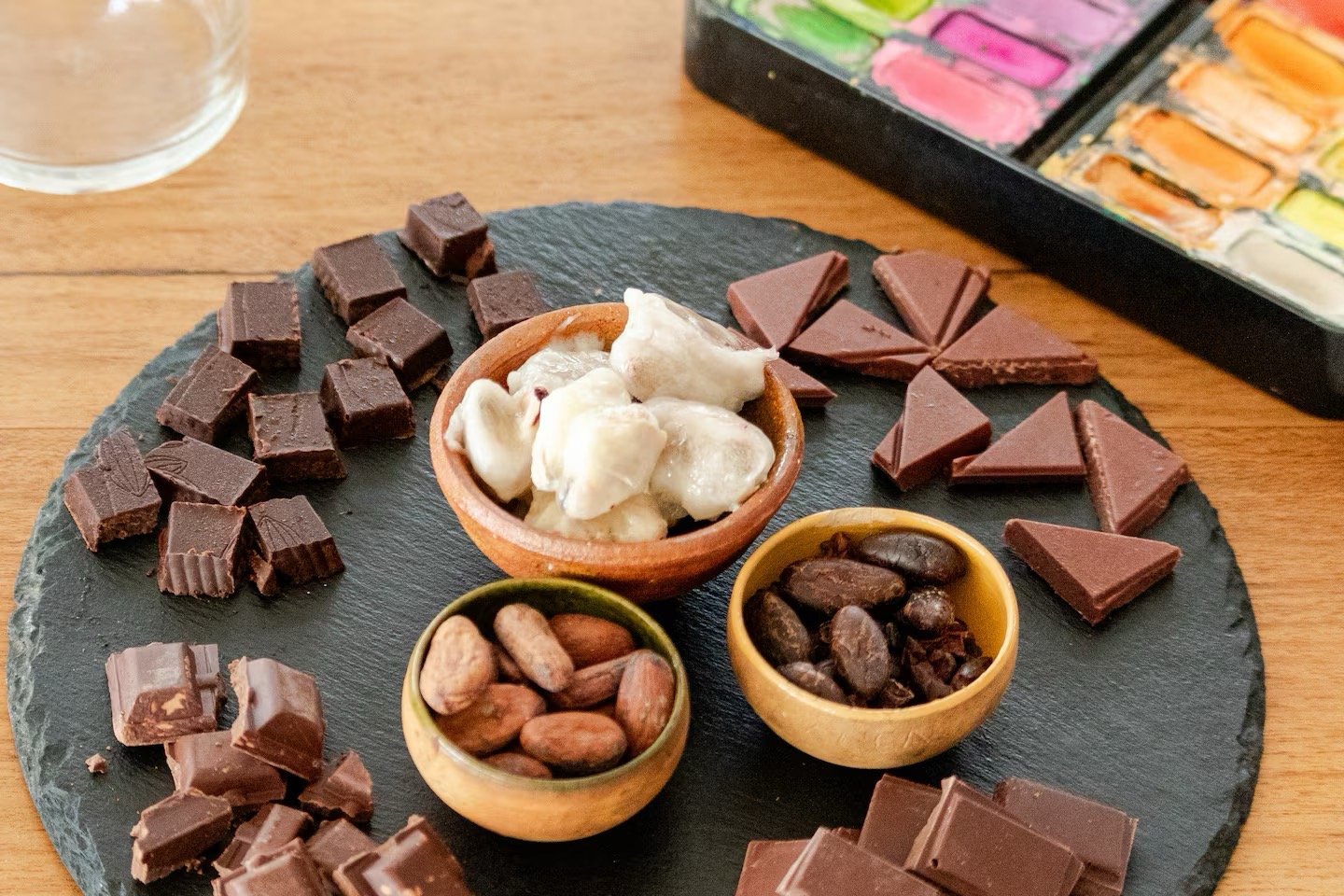 Assorted chocolates and nuts on a black slate platter with watercolor paints in the background.