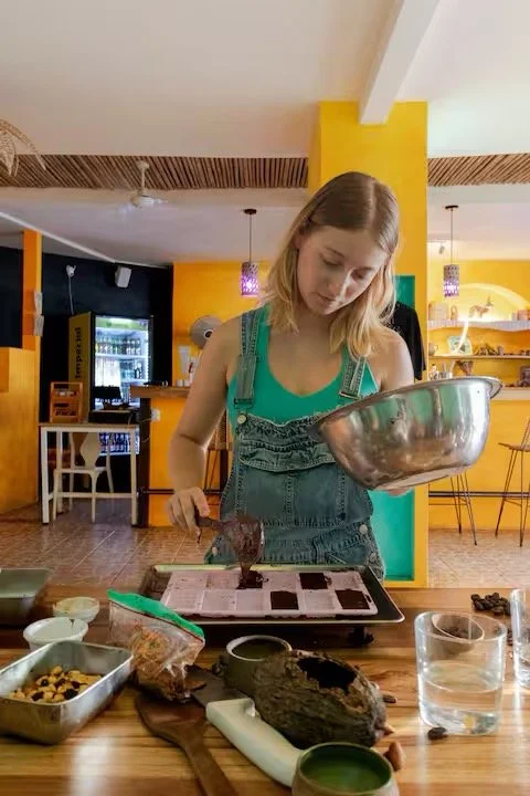 A young woman in overalls pouring chocolate batter into a baking tray in a colorful kitchen.