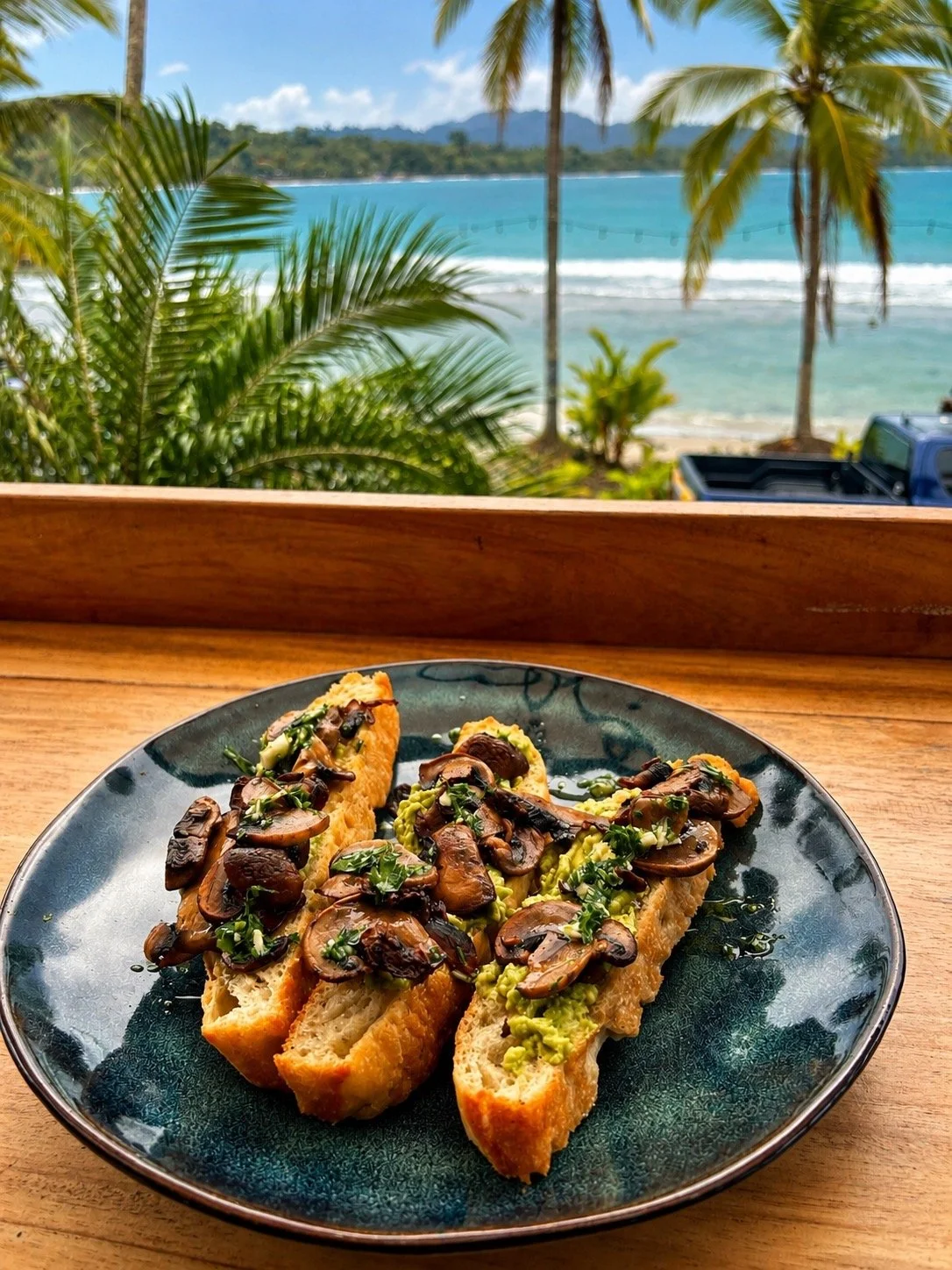 Three slices of toasted bread topped with sautéed mushrooms, chopped herbs, and guacamole, served on a dark plate against a beachside view with palm trees and the ocean in the background.