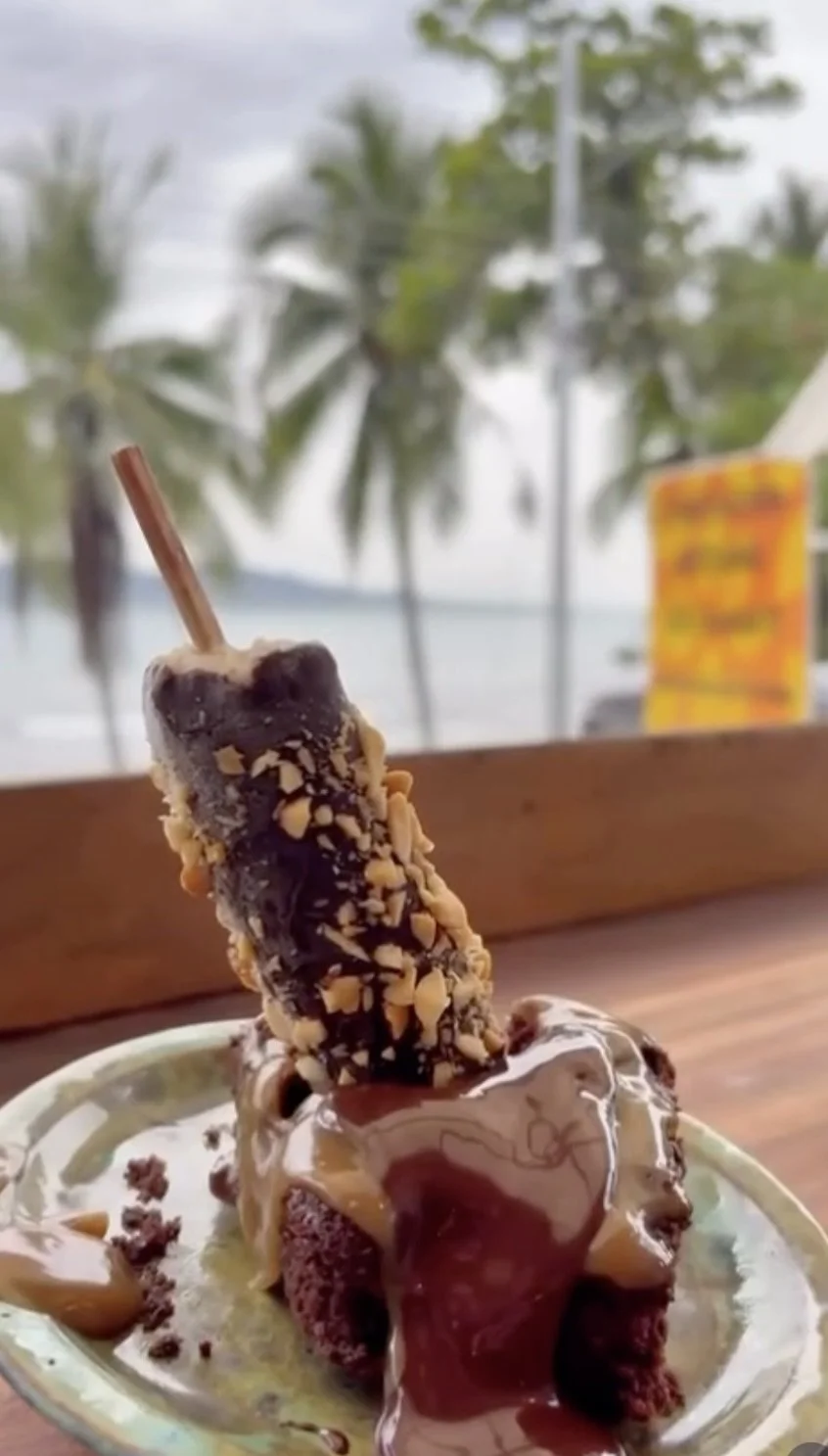 A vegan gluten free brownie with chocolate sauce, topped with a chocolate-covered frozen banana on a stick with chopped nuts, set against a background of palm trees and cloudy sky.