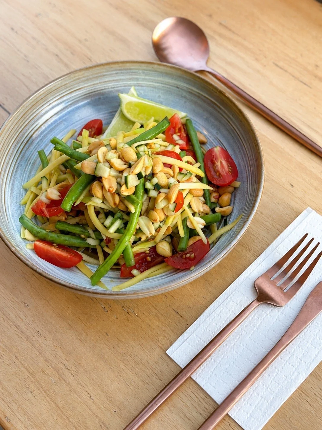 A bowl of salad containing cherry tomatoes, green beans, chopped cucumber, peanuts, and lime wedges on a wooden table, with a copper fork and spoon.