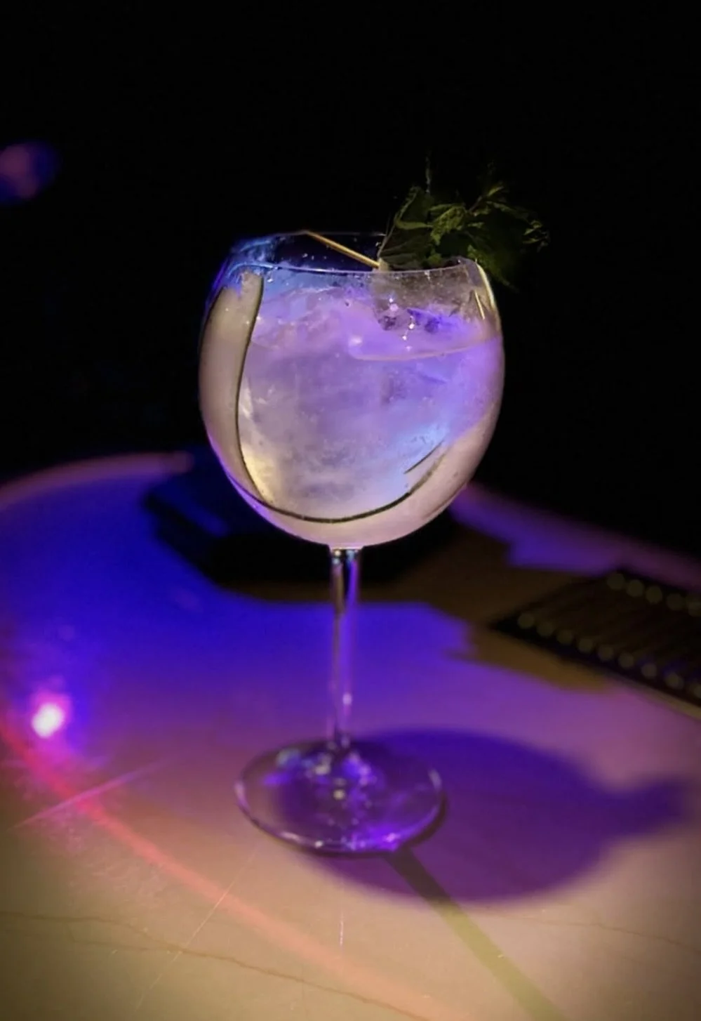 A cocktail with ice in a large wine glass, garnished with a sprig of mint, illuminated by purple and pink lighting.