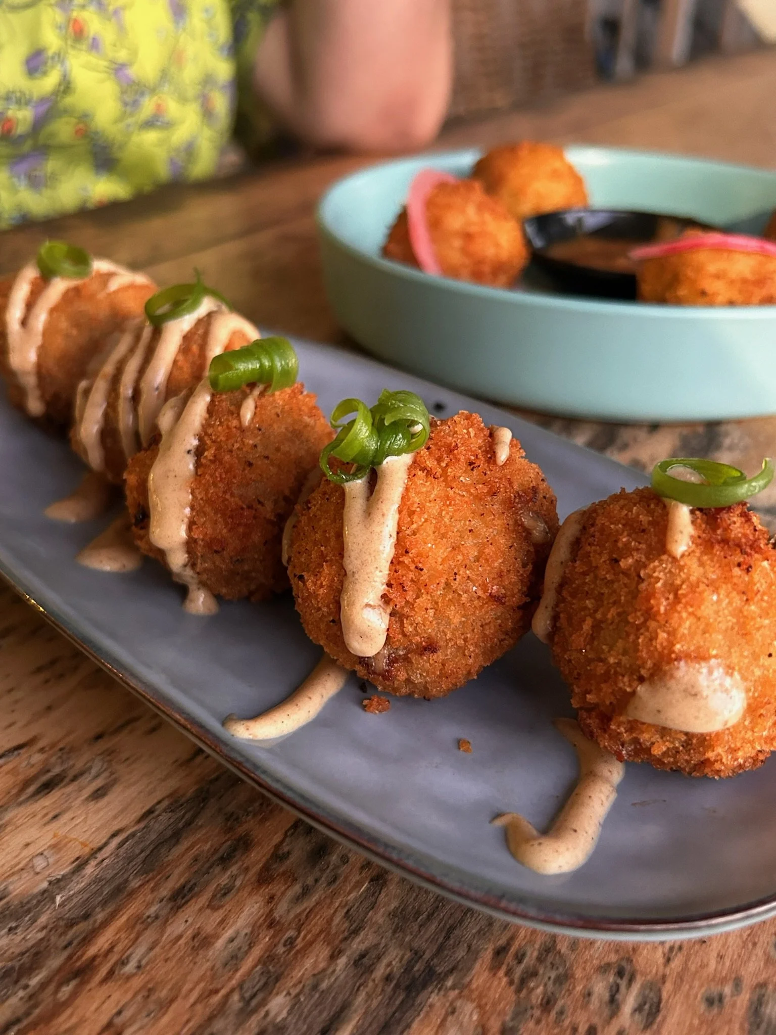 Close-up of Caribbean croquettes drizzled with a creamy sauce, garnished with chopped green onions, on a gray plate. In the background, there is a bowl with additional fried items and dipping sauce, on a wooden table.