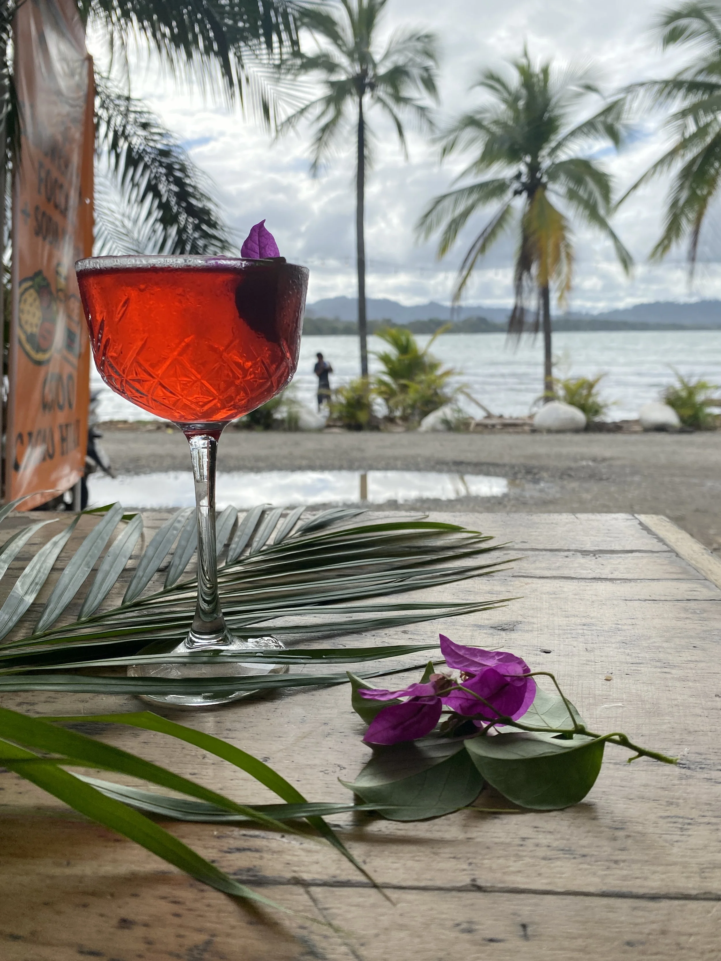 A hibiscus spritzer in a glass with a purple flower garnish sits on a table outdoors near water, with palm trees and mountains in the background.