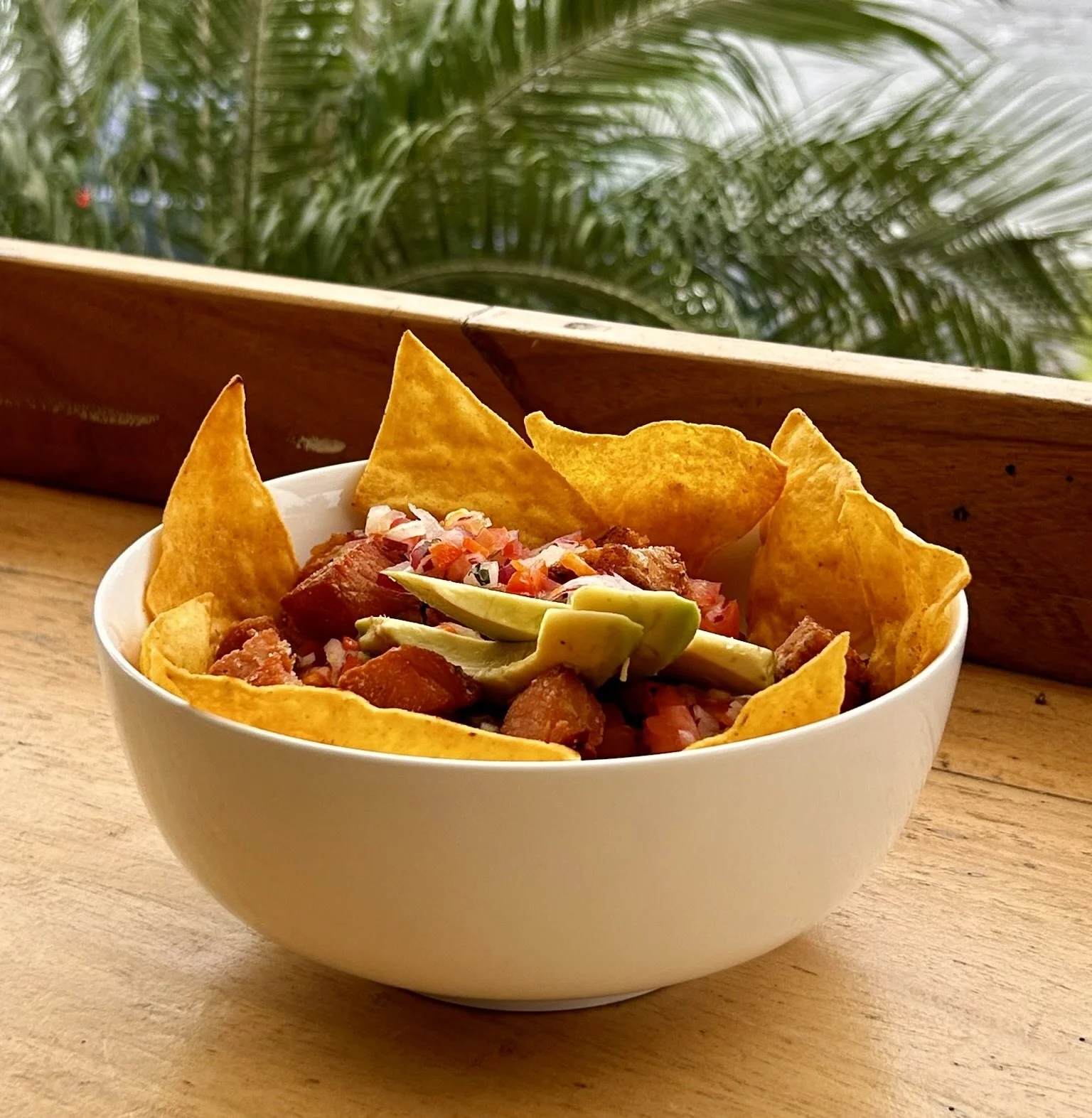 A white bowl filled with a Costa Rican Chifirjo, consisting of fried tortilla chips, shredded pork, sliced avocado, and a topping of diced tomatoes and onions, placed on a wooden table with plants in the background.