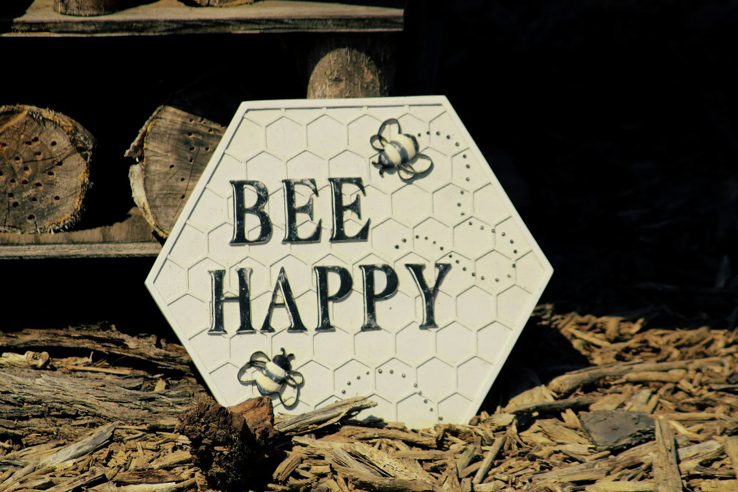 Woodpile with a white sign that says 'Bee Happy' and features a bee illustration. The sign is leaning against the woodpile, which is made of logs and bark chips.