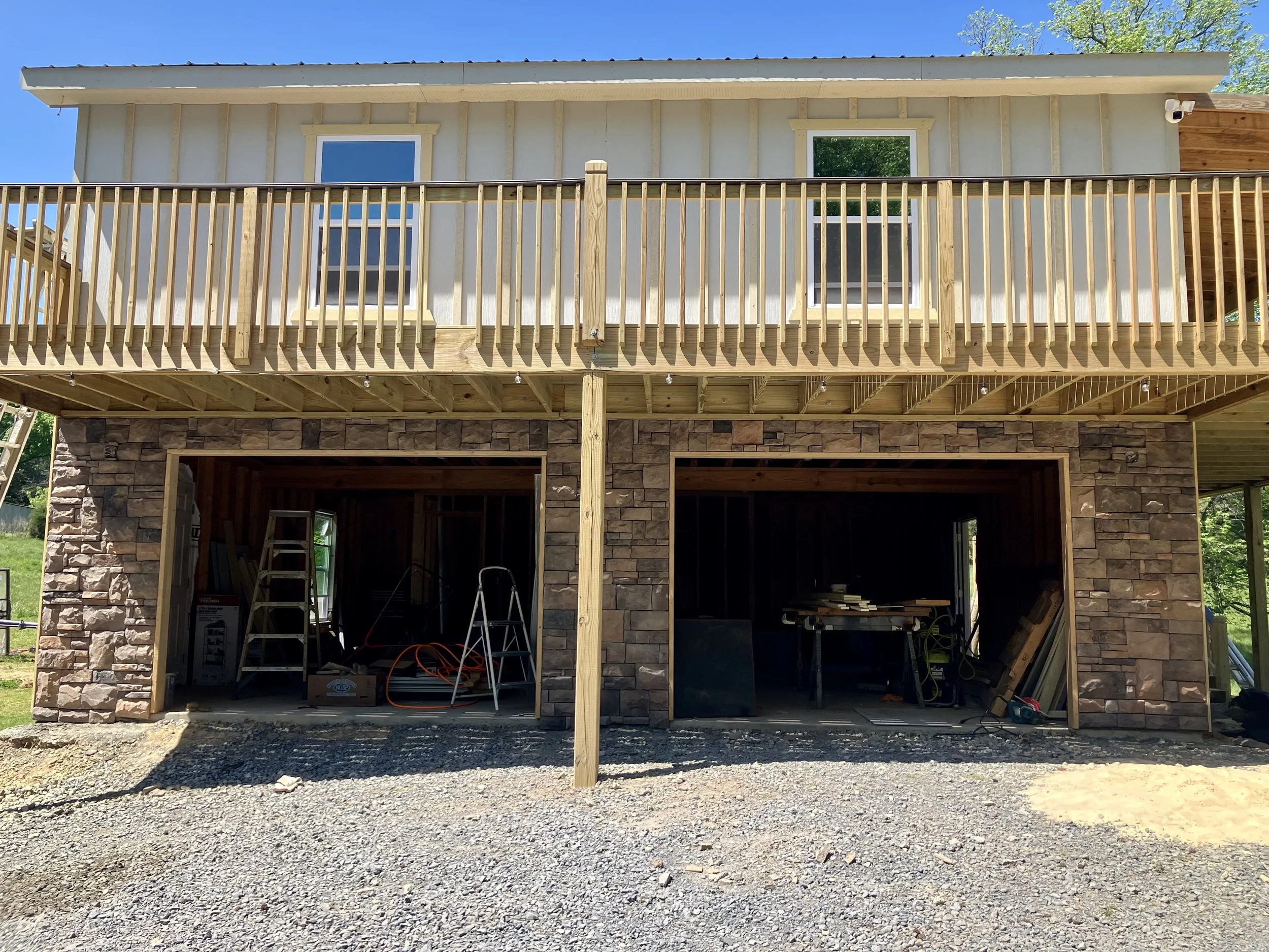Front view of a house under construction with partially completed stone walls to accent garage doors and a raised wooden deck with railing, supported by vertical posts; building materials and tools are inside the open garage area.