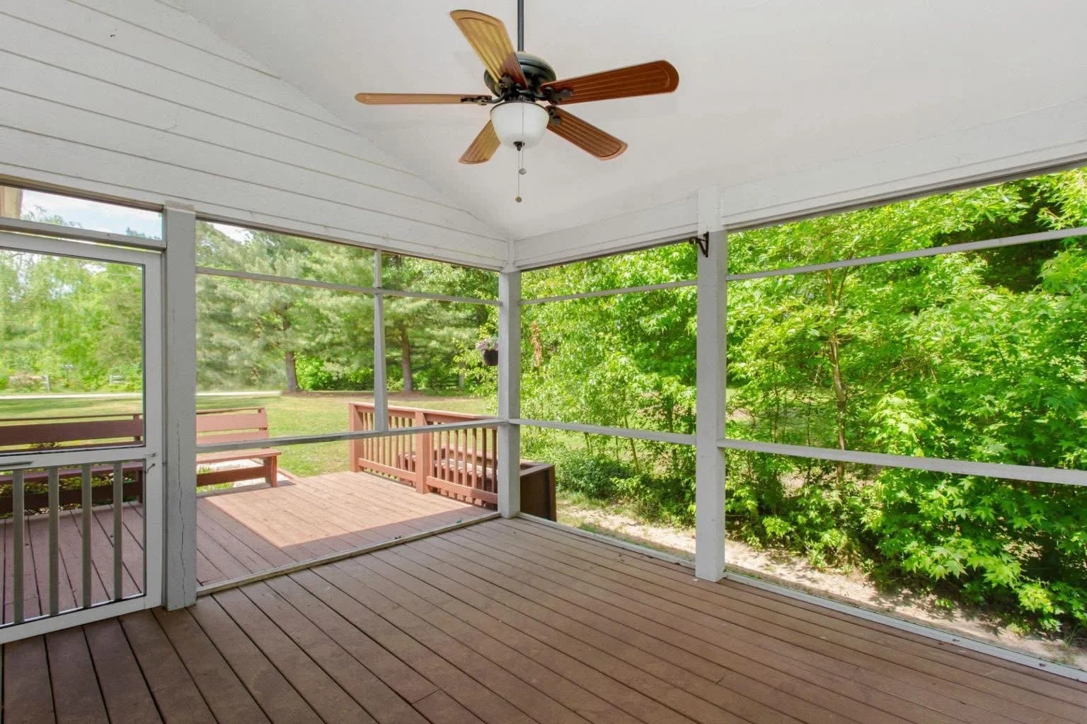 Enclosed porch with wooden floor and ceiling fan overlooking lush green trees and a neighboring deck with benches.