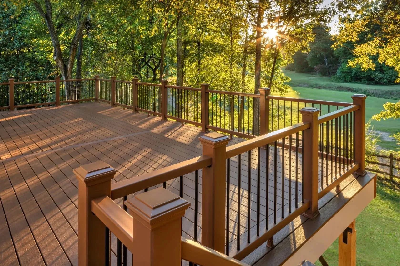 Wooden deck with black metal railing overlooking a lush green landscape with trees and a grassy field, illuminated by sunlight.