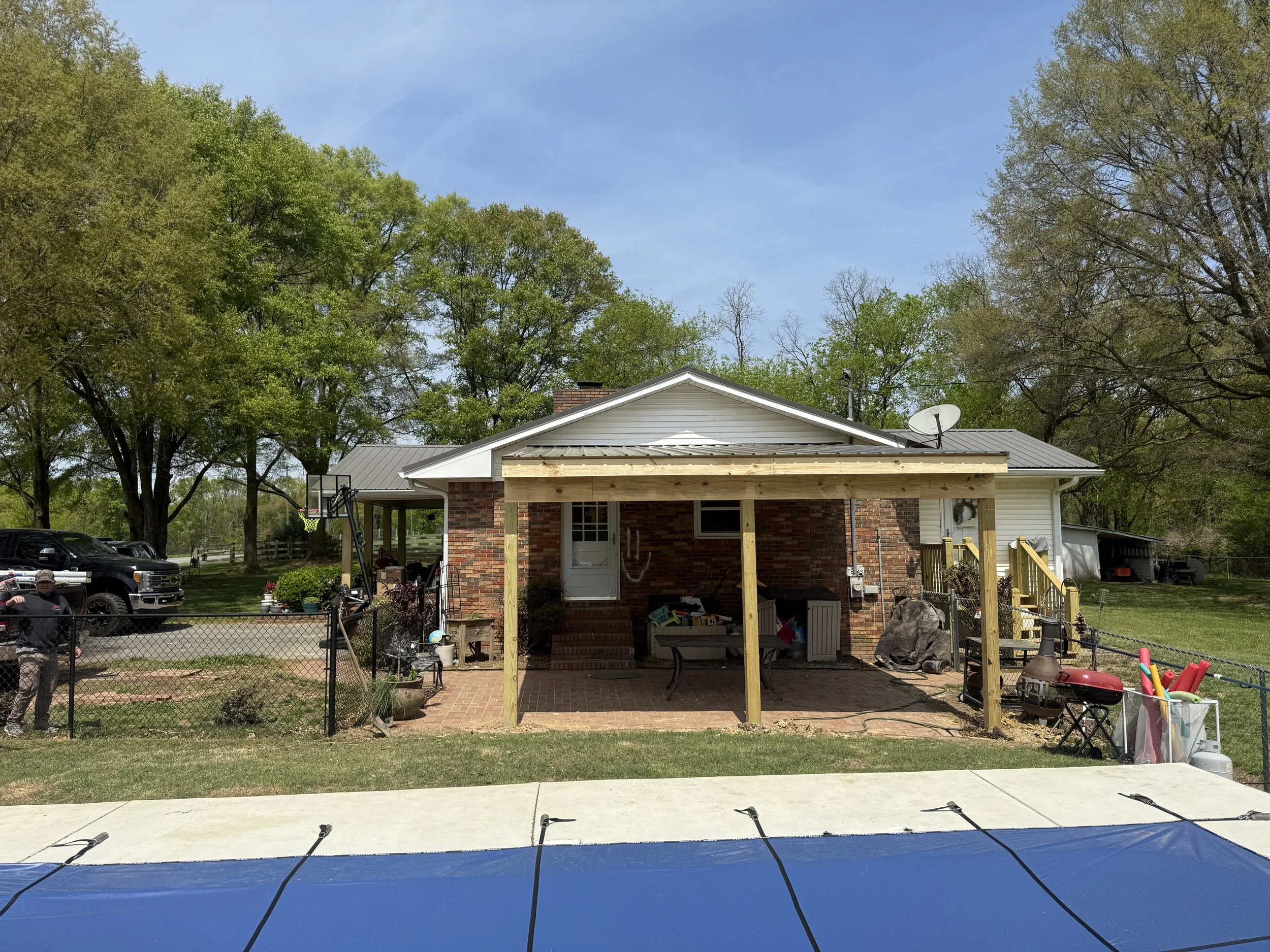 Front view of a house with a second-story porch in a backyard, surrounded by trees and a parking area with cars on the left. In the foreground, there is a trampoline with safety net.