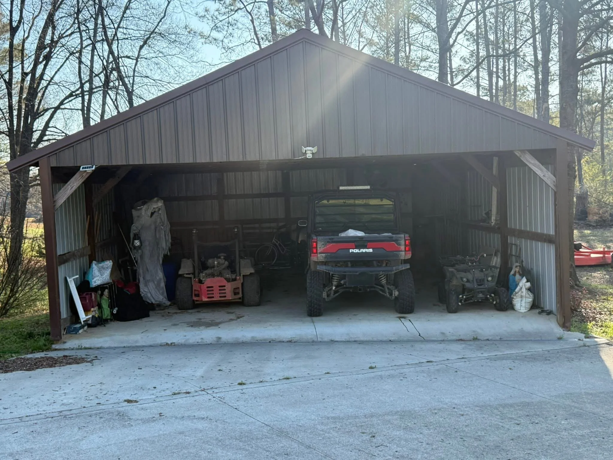 A spacious metal-sided garage with a peaked roof, open at the front, containing various outdoor equipment including a Polaris utility vehicle, bicycles, and gardening tools. The garage is situated in a wooded area with tall trees in the background an