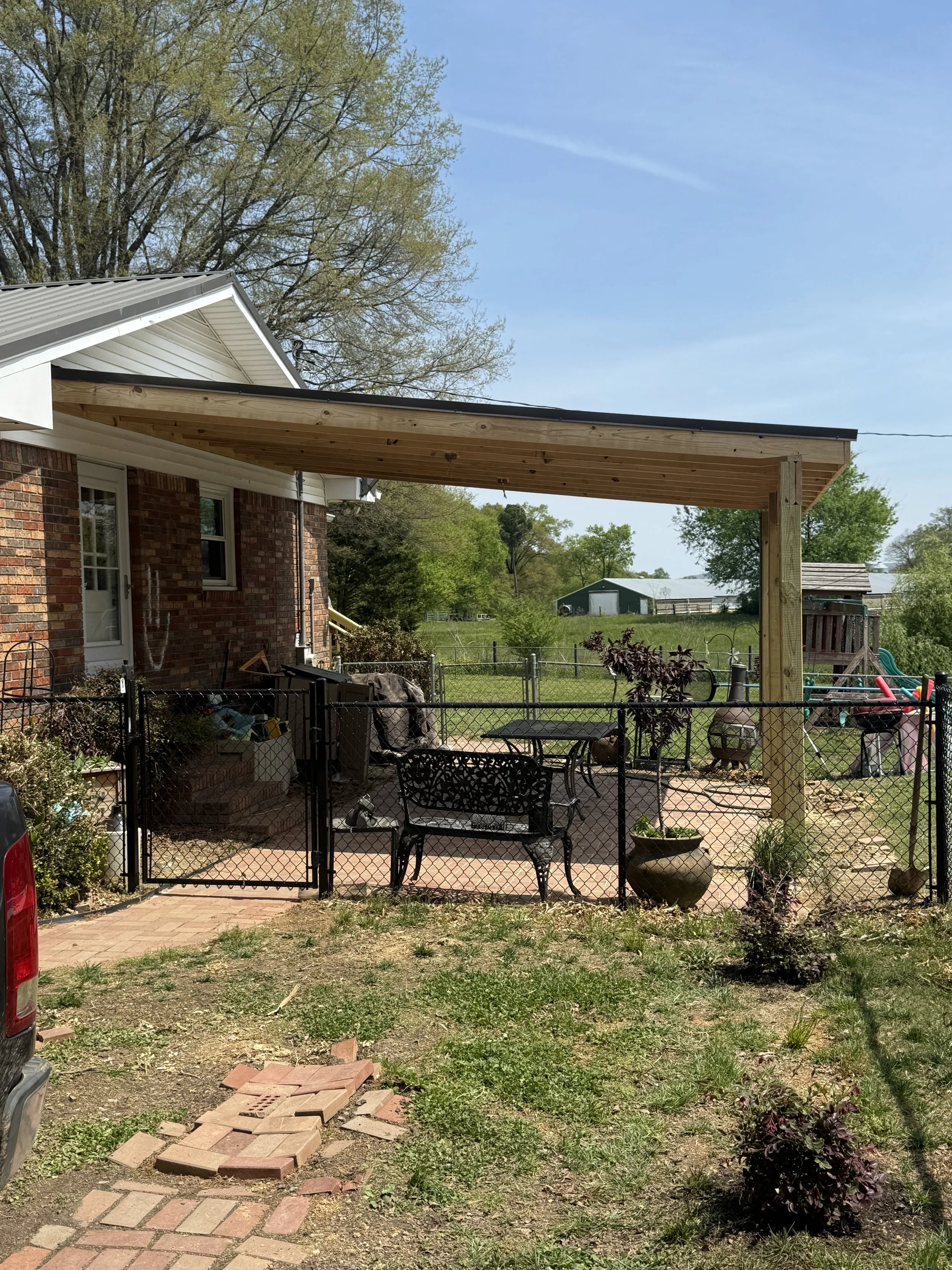 Backyard with brick house, metal roof, and wooden porch under construction. Fenced patio with outdoor furniture and potted plants, green grass, trees, and clear blue sky.