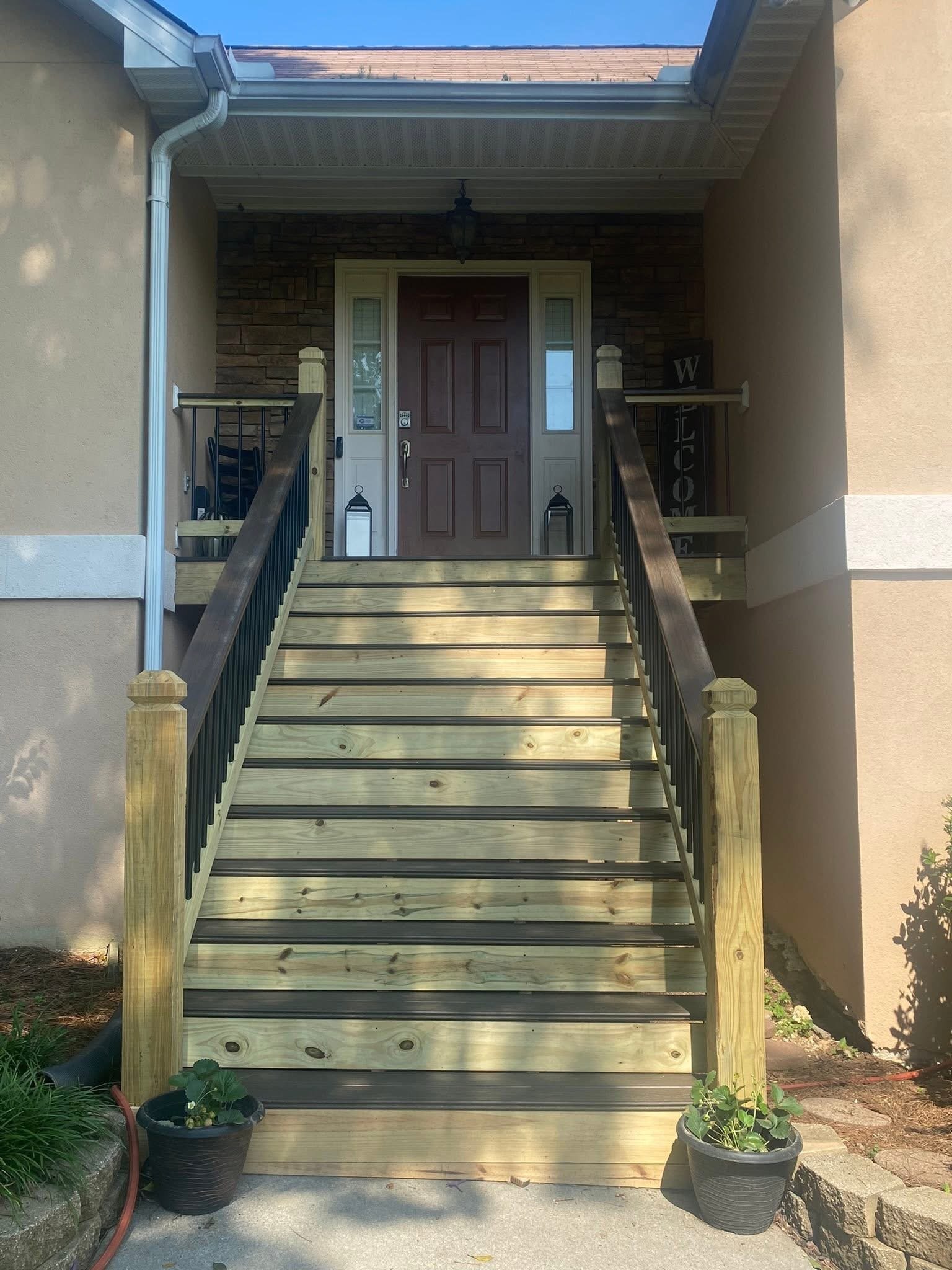 Front view of a house entrance with a trex board and wooden staircase, a brown door, two potted plants on the ground, and a small roof with gutter system.