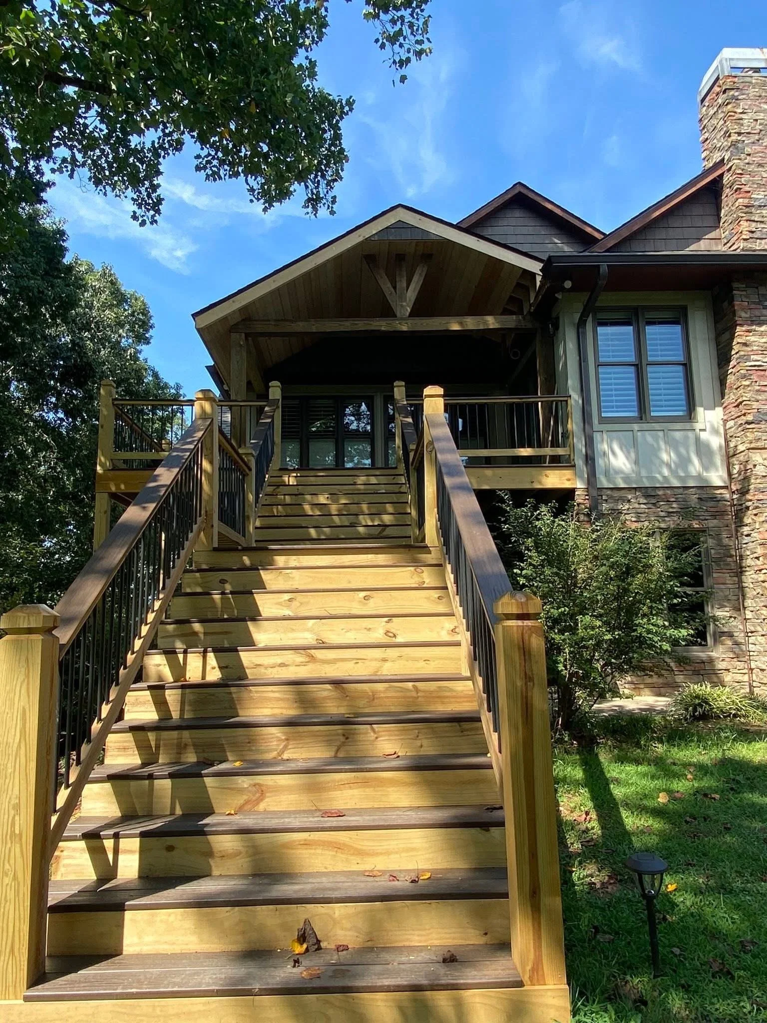 Newly built wooden staircase with black metal railing leading up to a house with a covered porch. The house has a brick chimney, large windows with white shutters, and a modern design with wood and stone elements. Green trees and grass surround the h