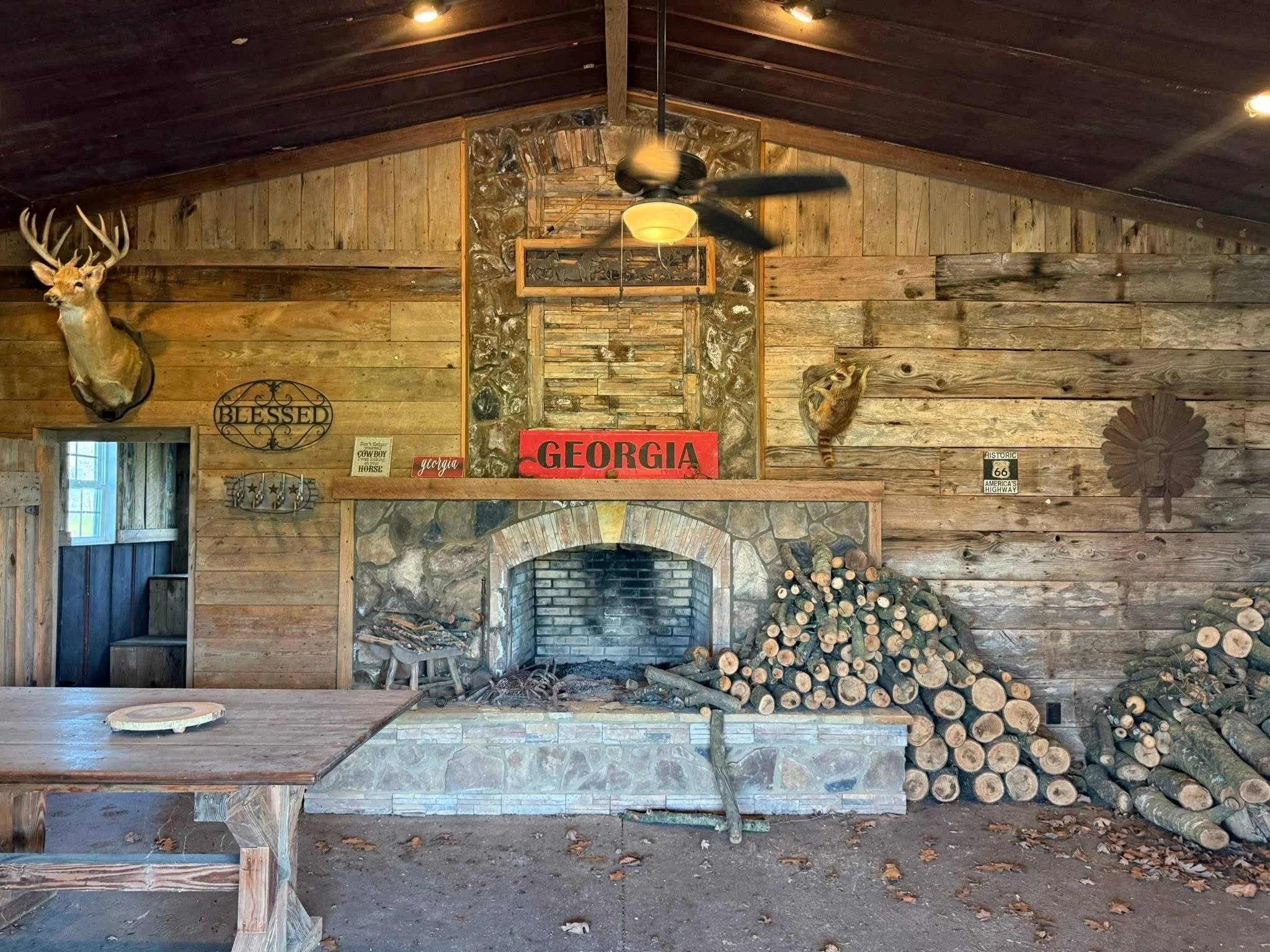 A rustic pavillion with wooden walls and ceiling, entertaining area, stone fireplace, a ceiling fan with lights, a private bathroom, sink, and plumbing.