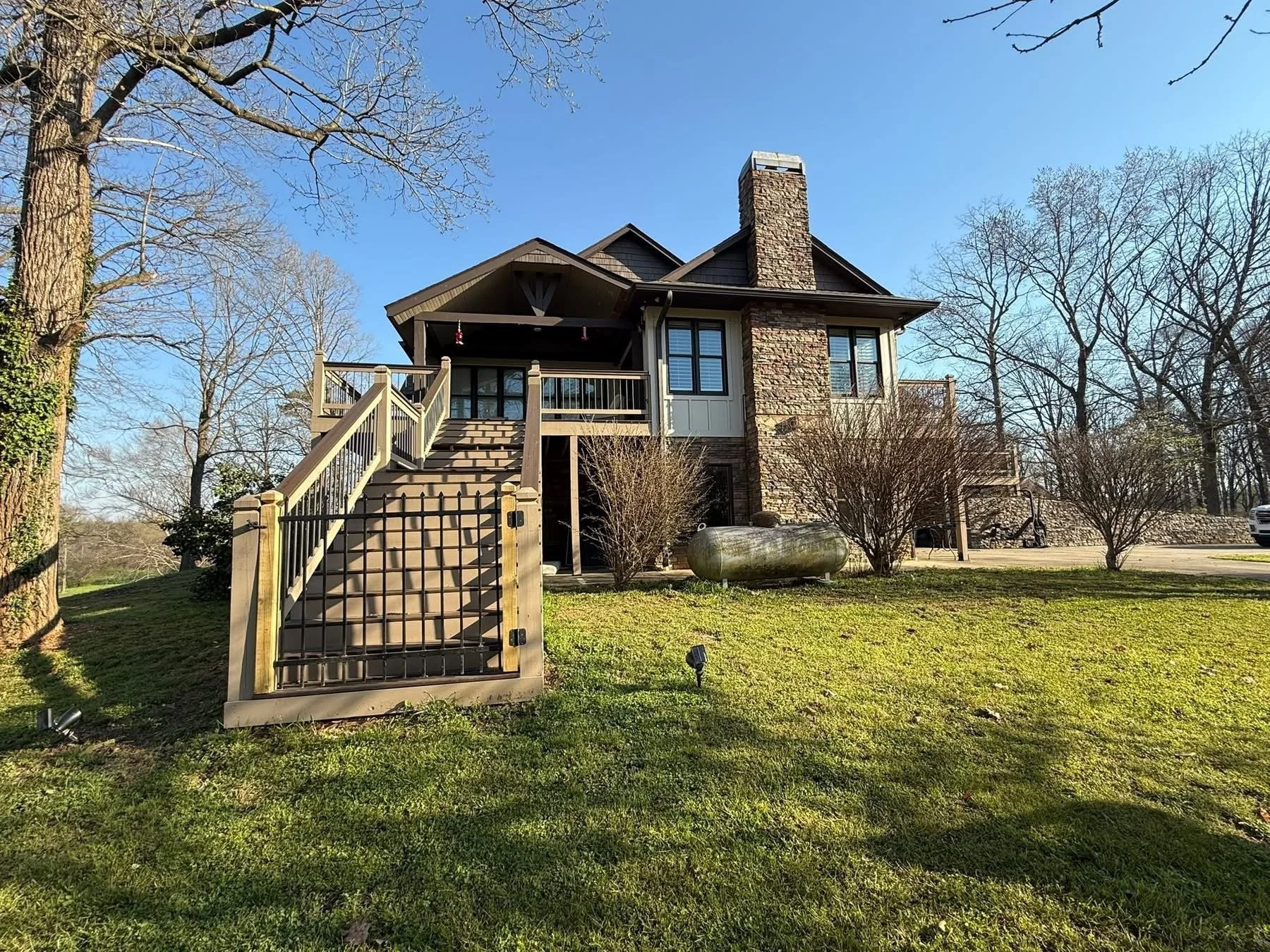 A two-story house with a stone chimney, a wooden staircase leading to a balcony, surrounded by trees and a grassy yard with outdoor lighting fixtures.