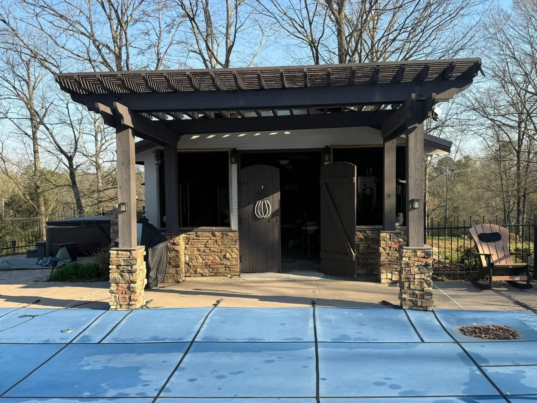 Backyard with a covered patio, house with open black doors, a swimming pool covered with a safety cover, leafless trees, outdoor chairs, and various items near the house.